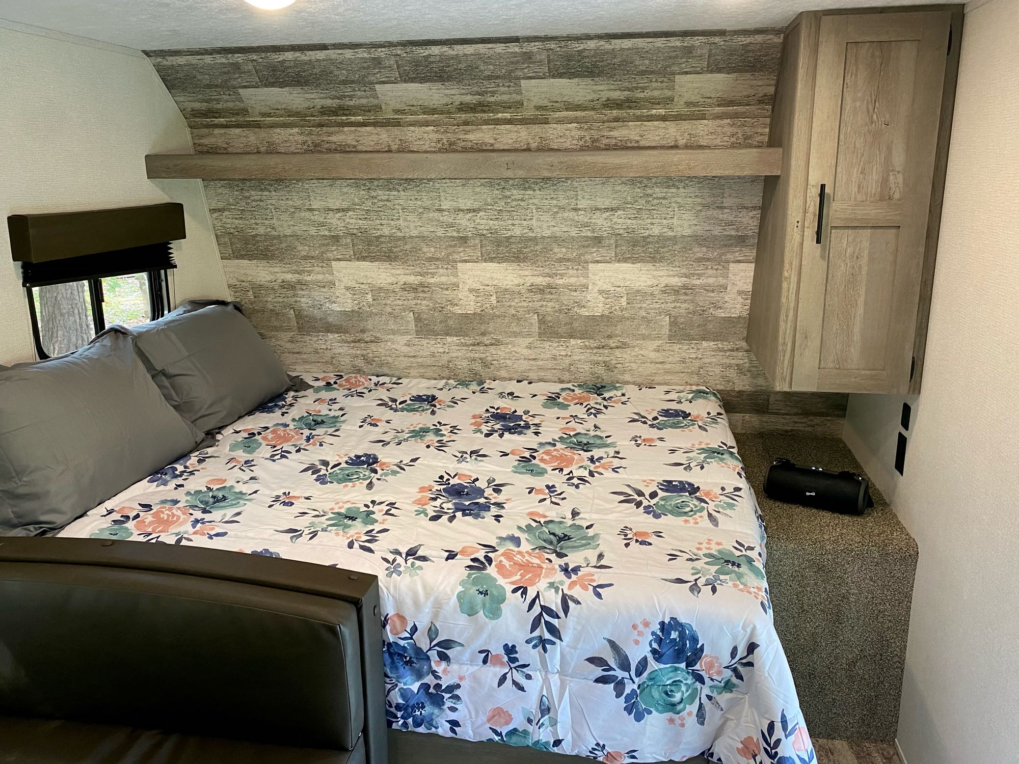 RV rental bedroom: a cozy space with a floral comforter on the bed, grey pillows, wood accent wall, cabinet, and window.. Keystone RV Springdale 2021