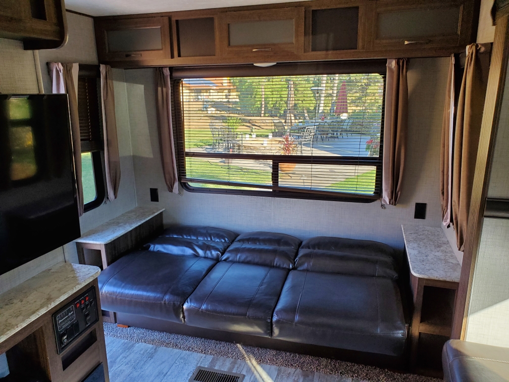 Inside an **RV rental**: a brown leather sofa, TV, and large window overlooking an outdoor patio scene.. Crossroads Zinger 2018
