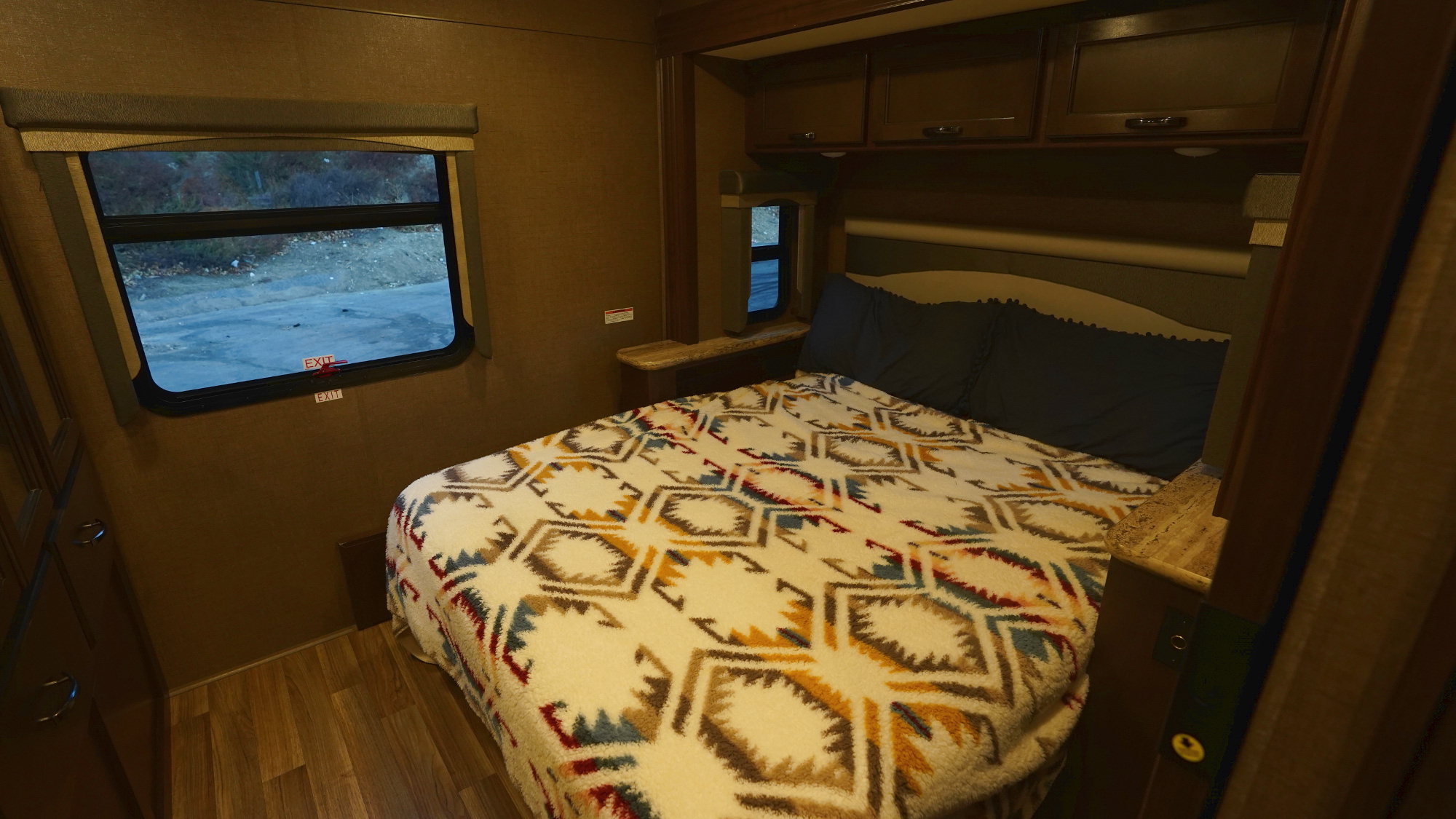 Cozy RV rental bedroom showing a bed with a patterned blanket, dark pillows, window view, and wooden cabinetry.. Thor Motor Coach A.C.E 2020