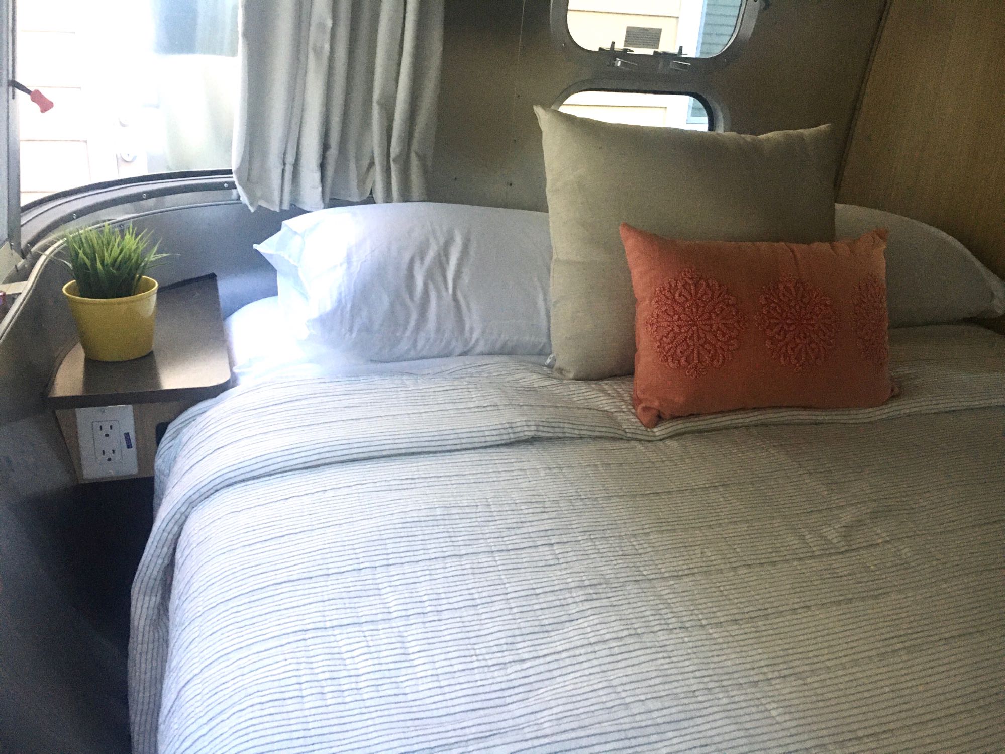 Cozy Airstream RV rental bedroom interior with comfortable bed, striped bedding, and plant. Ideal for travel accommodation.. Airstream Flying Cloud 2019