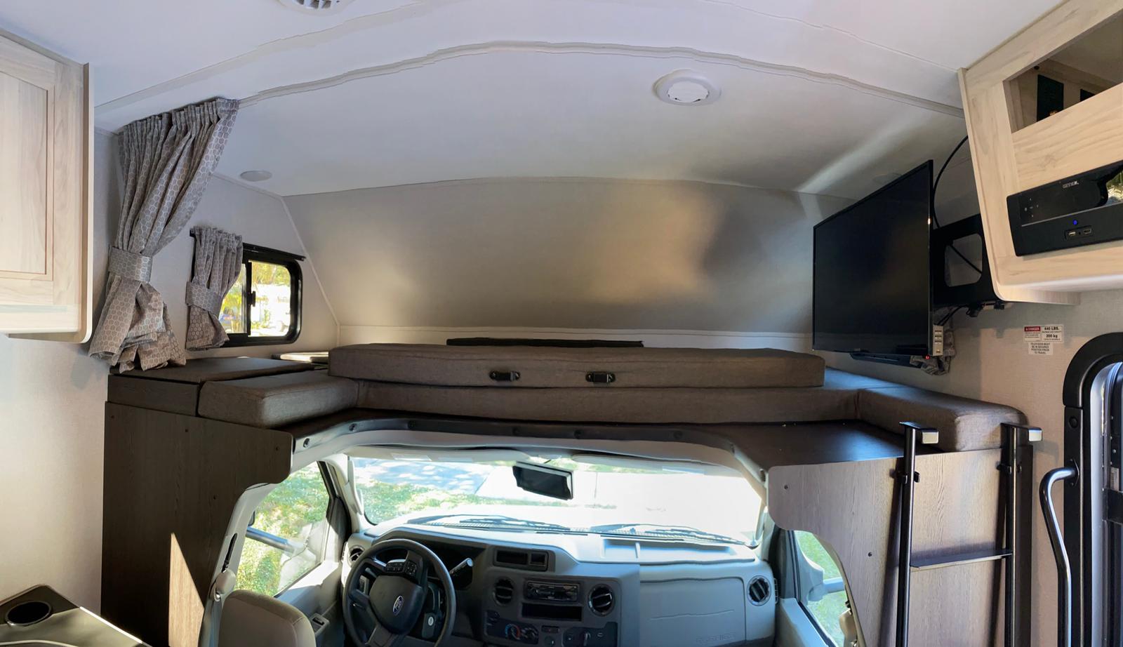 View from the living area towards the front of an RV rental, showing the driver's cab under the overhead bunk with TV.. Forest River Forester 2021