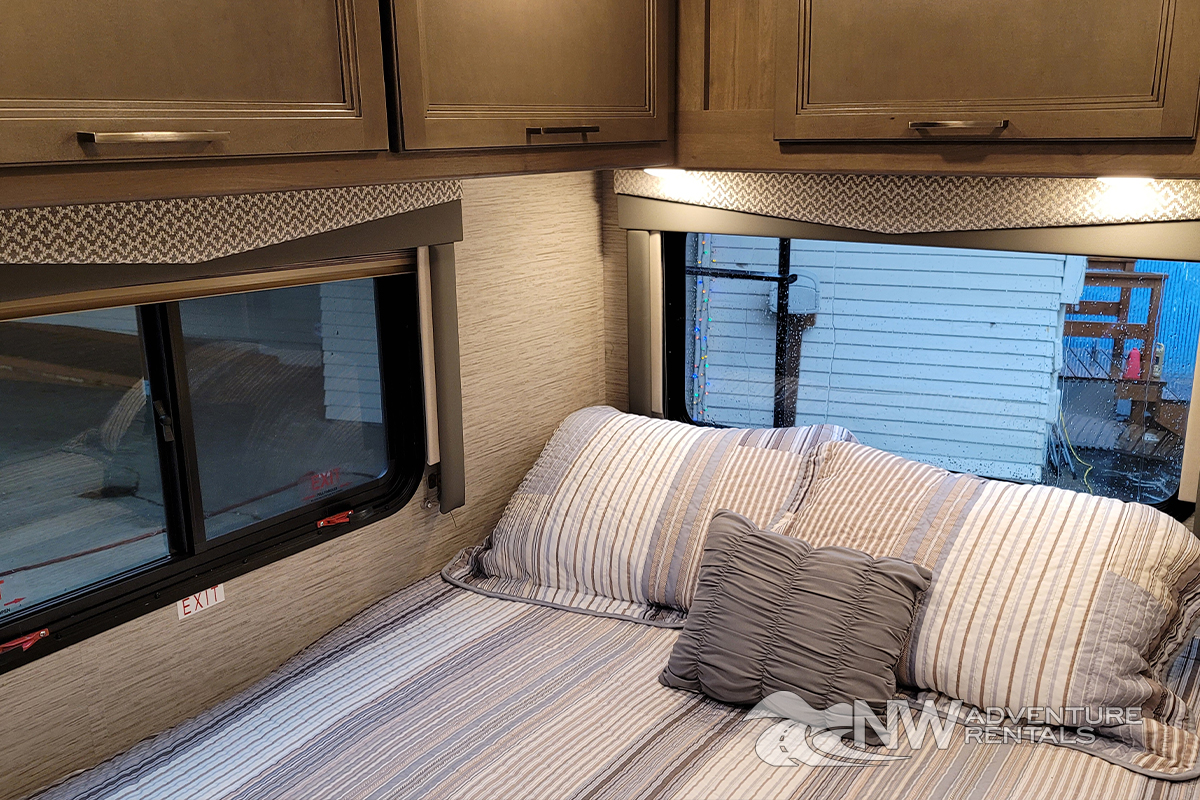 Cozy RV rental bedroom with comfortable striped bed, overhead cabinets, and bright windows.. Thor Motor Coach Four Winds 2021