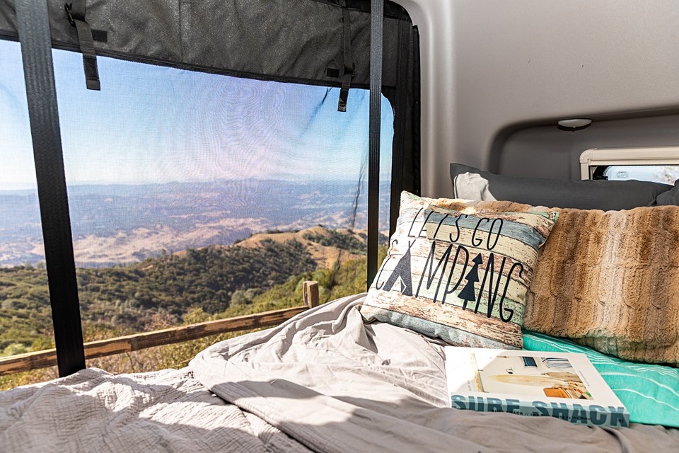 An RV rental bed, with a 'LET'S GO CAMPING' pillow, offers sweeping mountain views through a mesh screen.. Winnebago Revel 2021