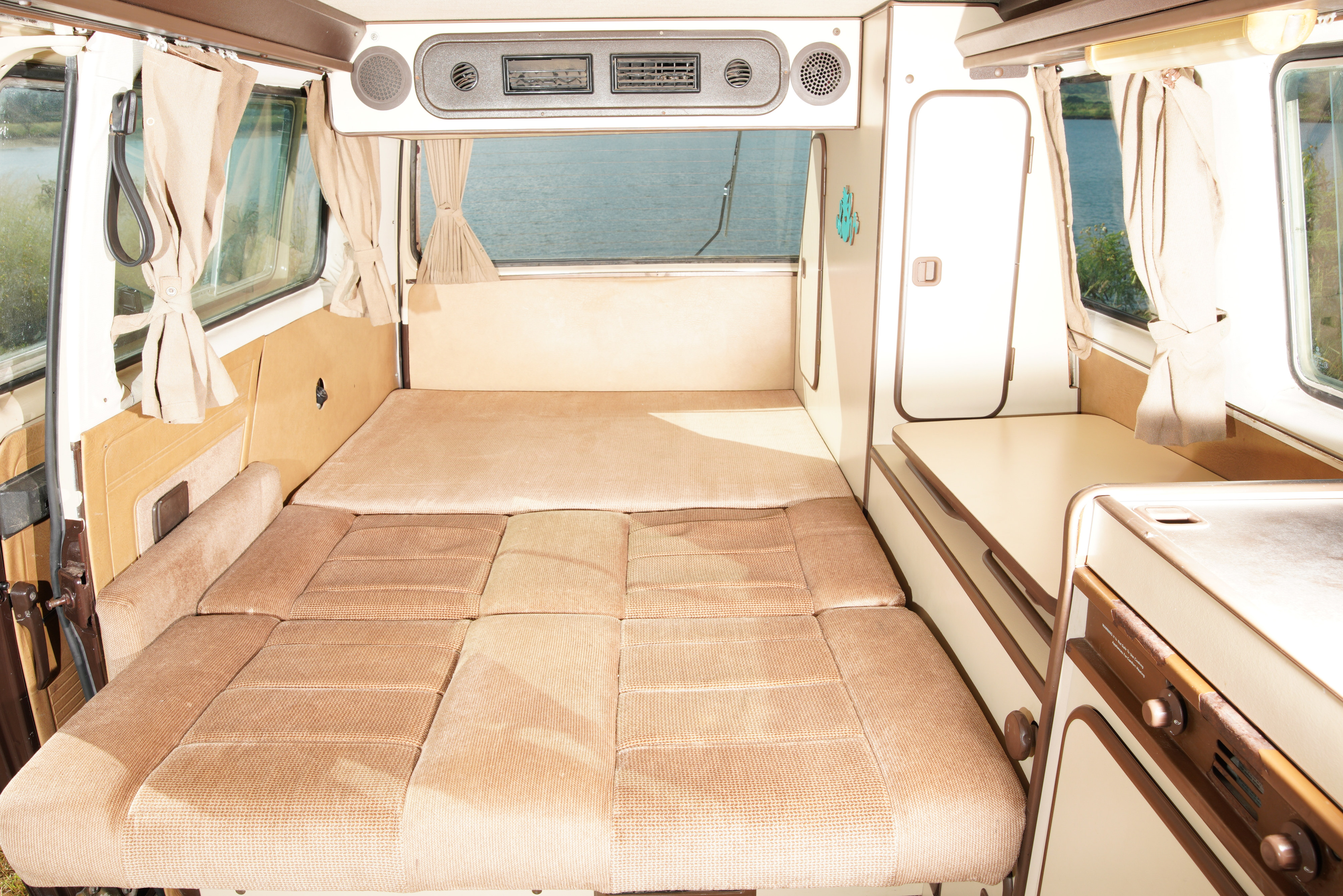 Cozy RV rental interior featuring a convertible bed, compact kitchenette, and windows revealing a scenic lake view.. Volkswagen Westfalia 1985