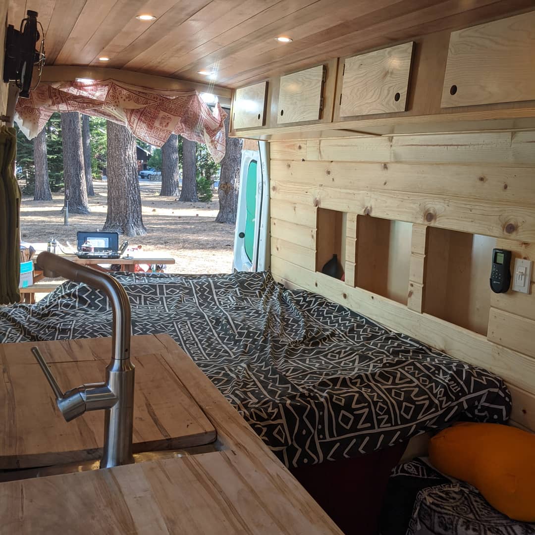 Inside a wooden rv rental camper van: built-in kitchen, comfy bed, and a scenic forest view out the back. Ideal for van life.. Ford Transit Custom 2019