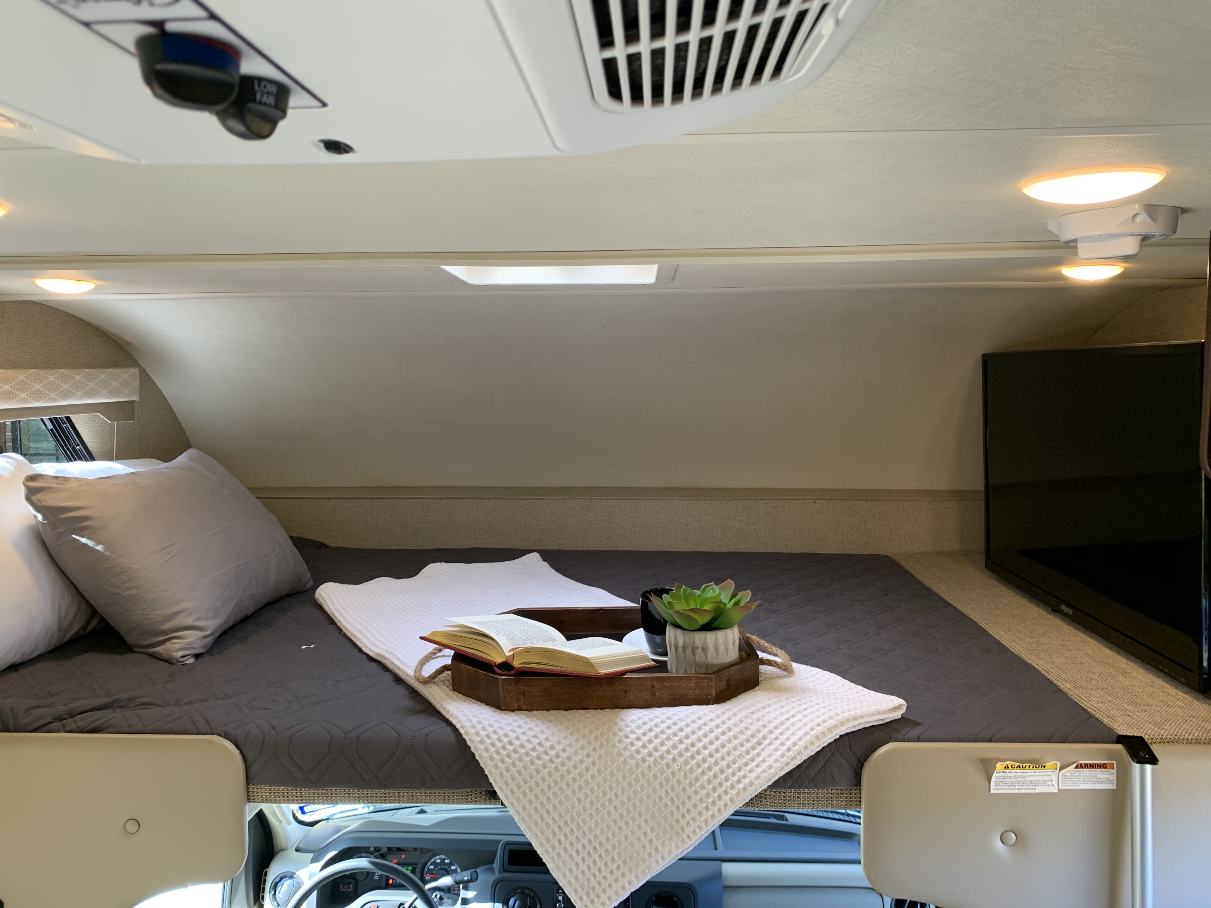 Cozy RV rental cab-over bunk features a bed, TV, and thoughtful comforts like a book and plant.. Thor Freedom Elite 2019