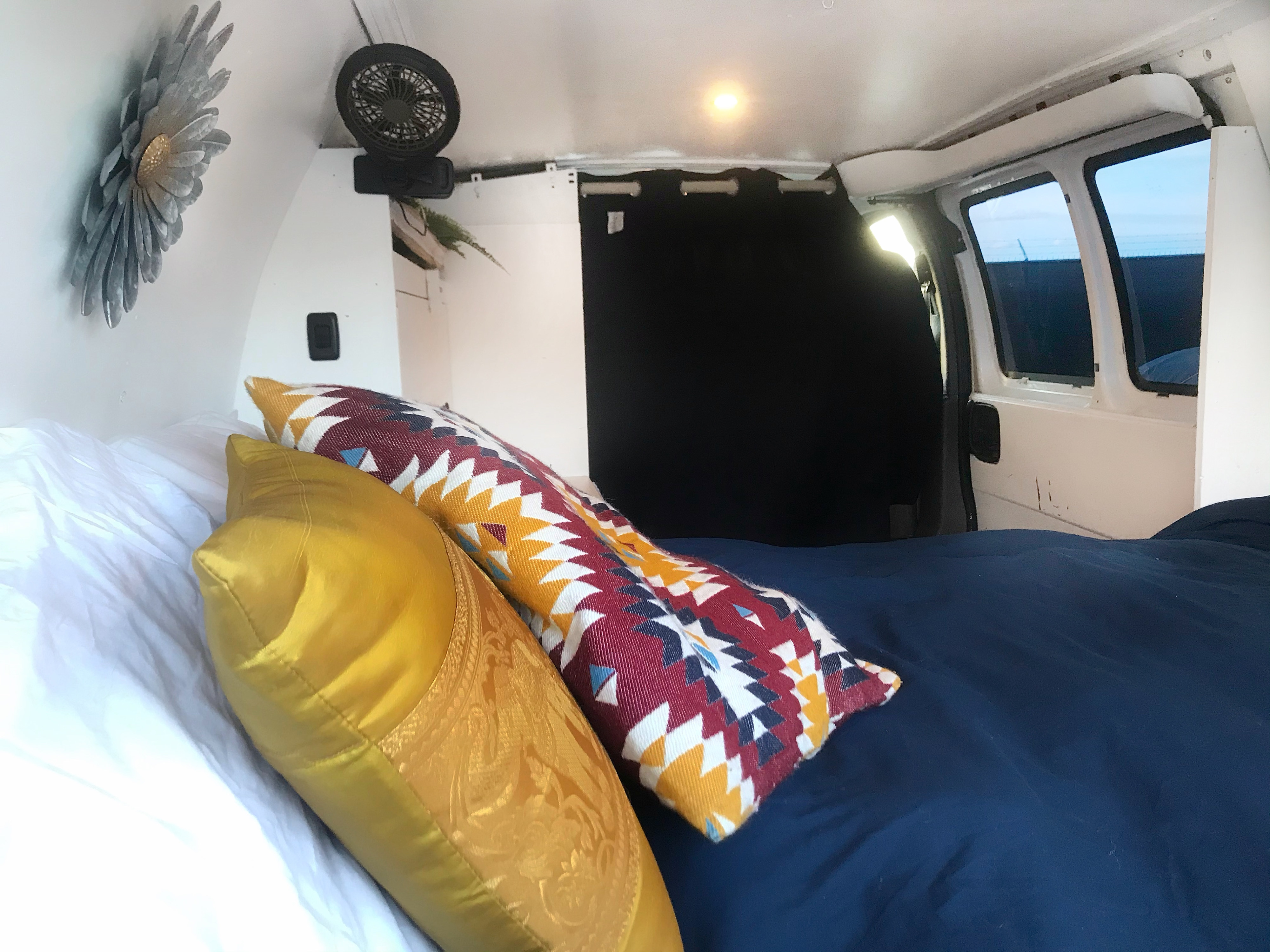 Cozy RV rental interior with a comfortable bed, decorative pillows, fan, and rear windows. Ideal for van life adventures.. Chevrolet Express 1500 2006