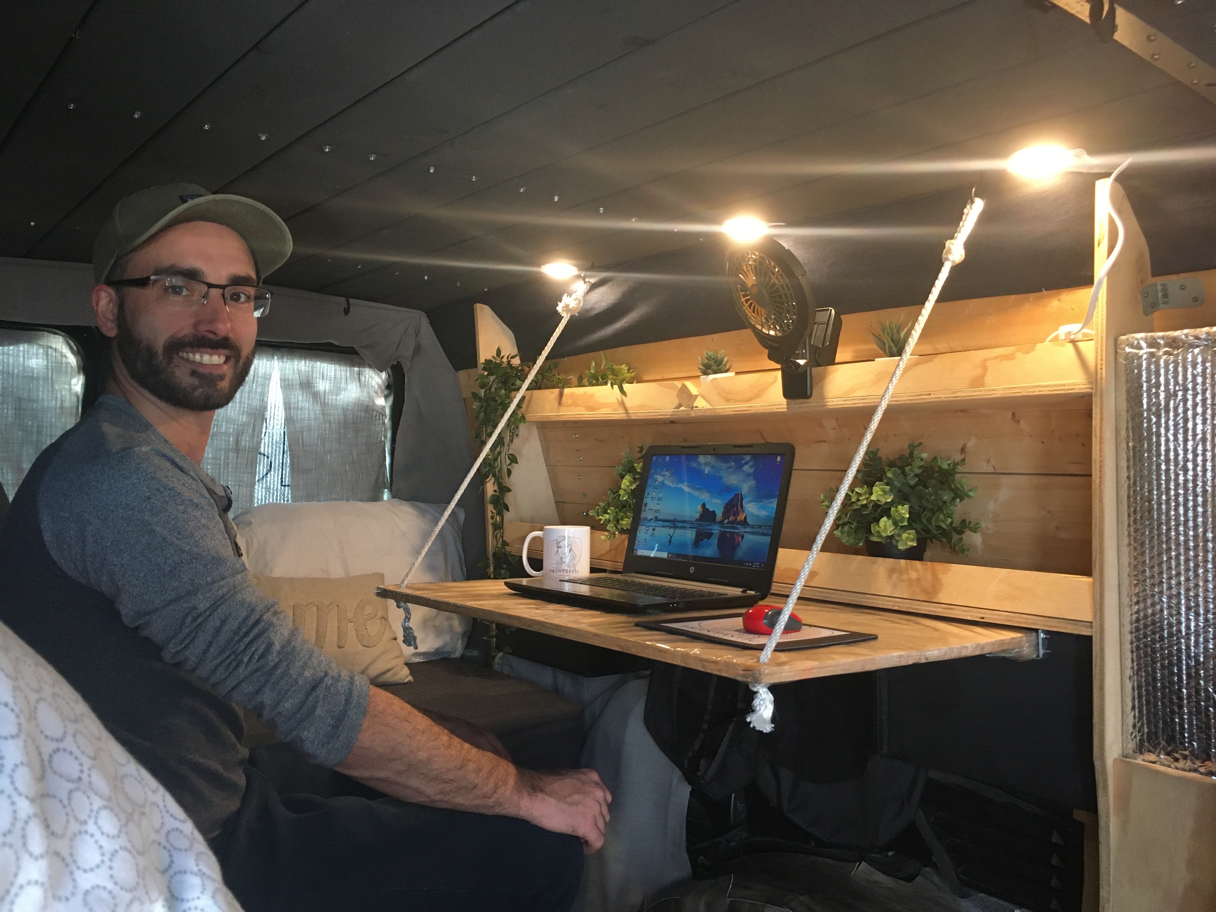 A man smiles inside a cozy RV rental workspace, featuring a laptop on a suspended desk, ambient lights, and plants.. Ford Econoline Extended 2006