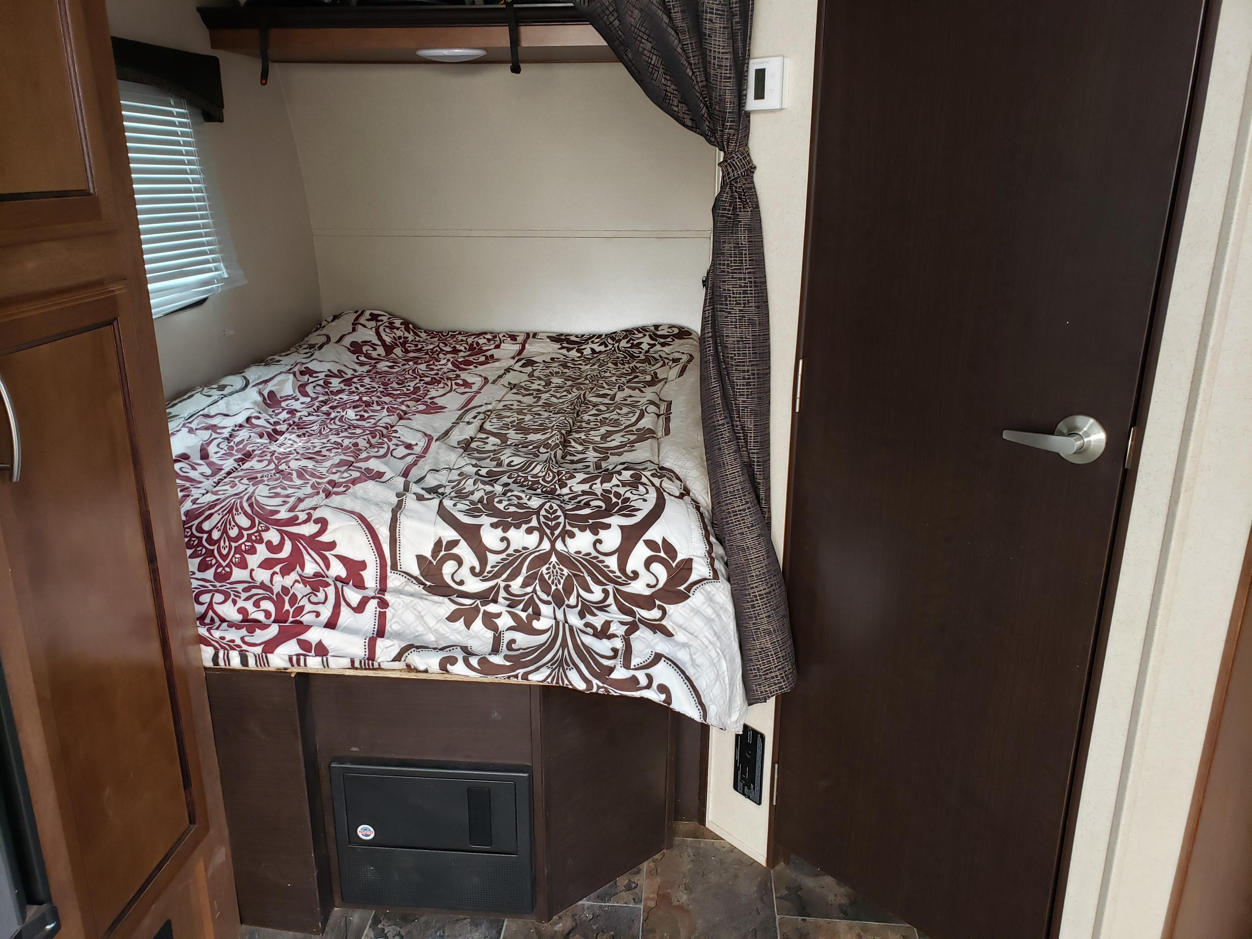 Compact RV rental bedroom showing a patterned bed, window, dark curtain, thermostat, and brown interior door.. Forest River Shockwave 2017