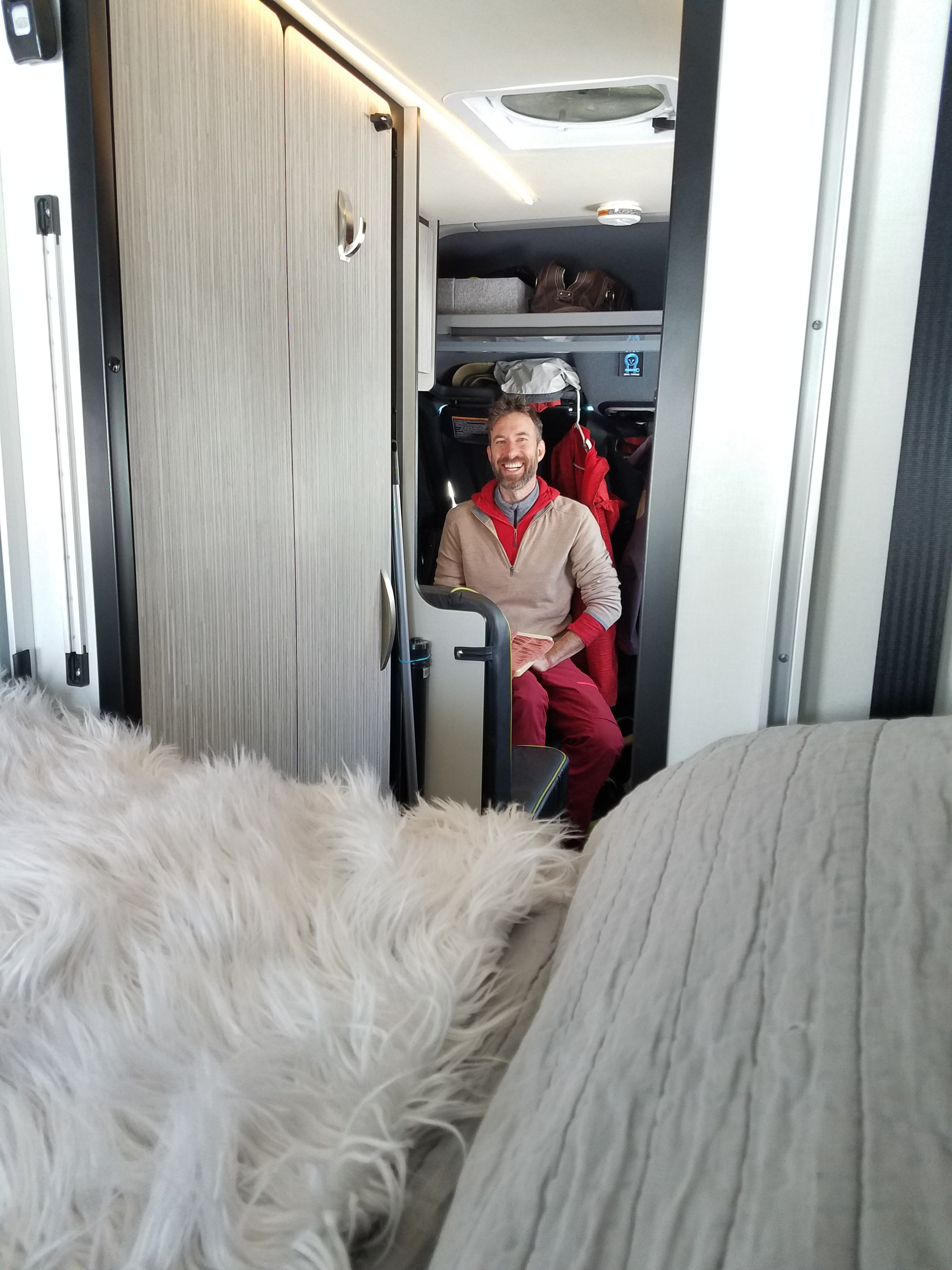 Happy man inside a comfy RV rental, showcasing the cozy living area with storage and a fluffy blanket.. Winnebago Revel 2020