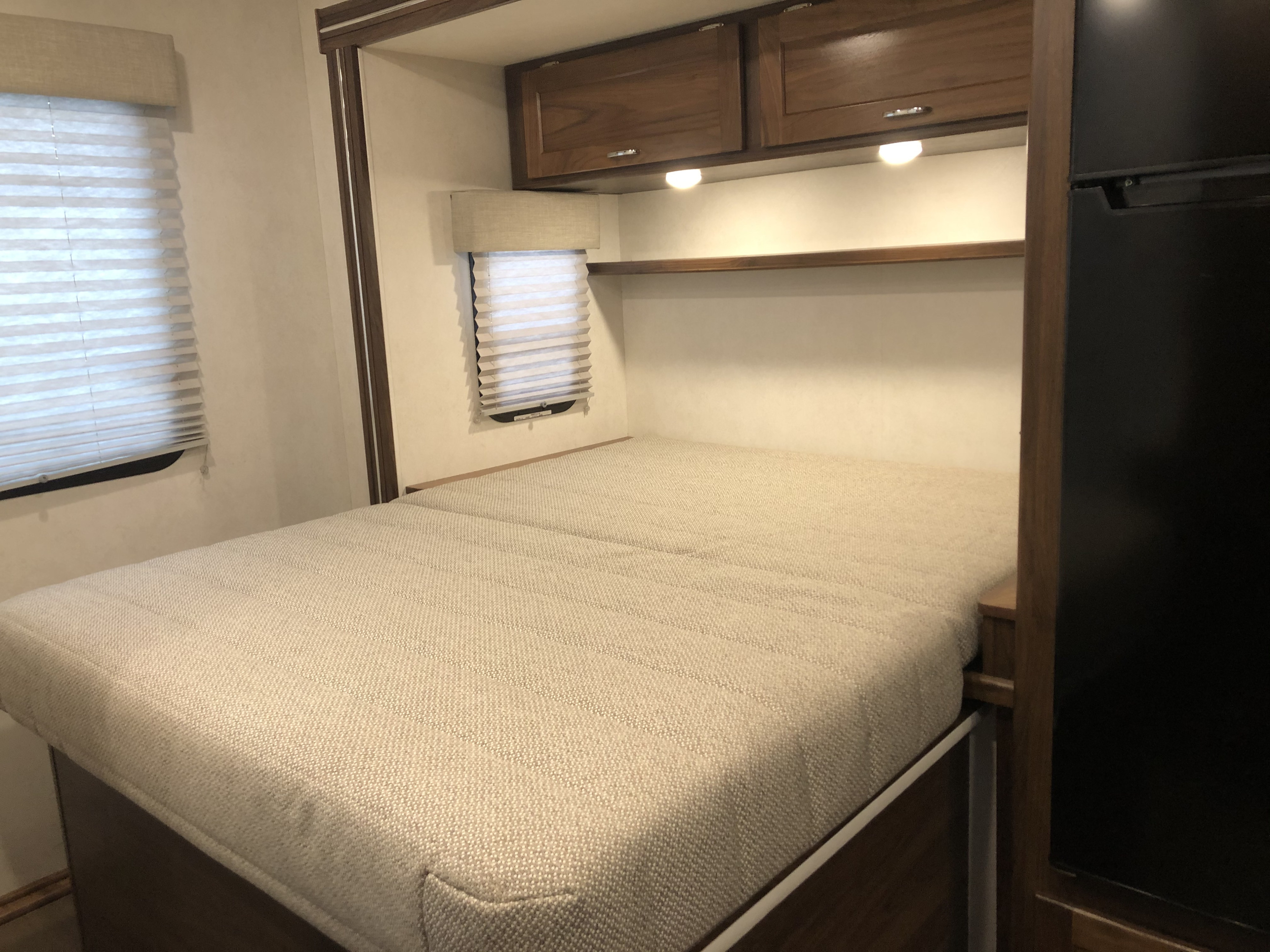 RV rental sleeping area: light, textured bed, wood cabinets with lights, cellular blinds, and a black refrigerator.. Winnebago Mercedes Sprinter 2020