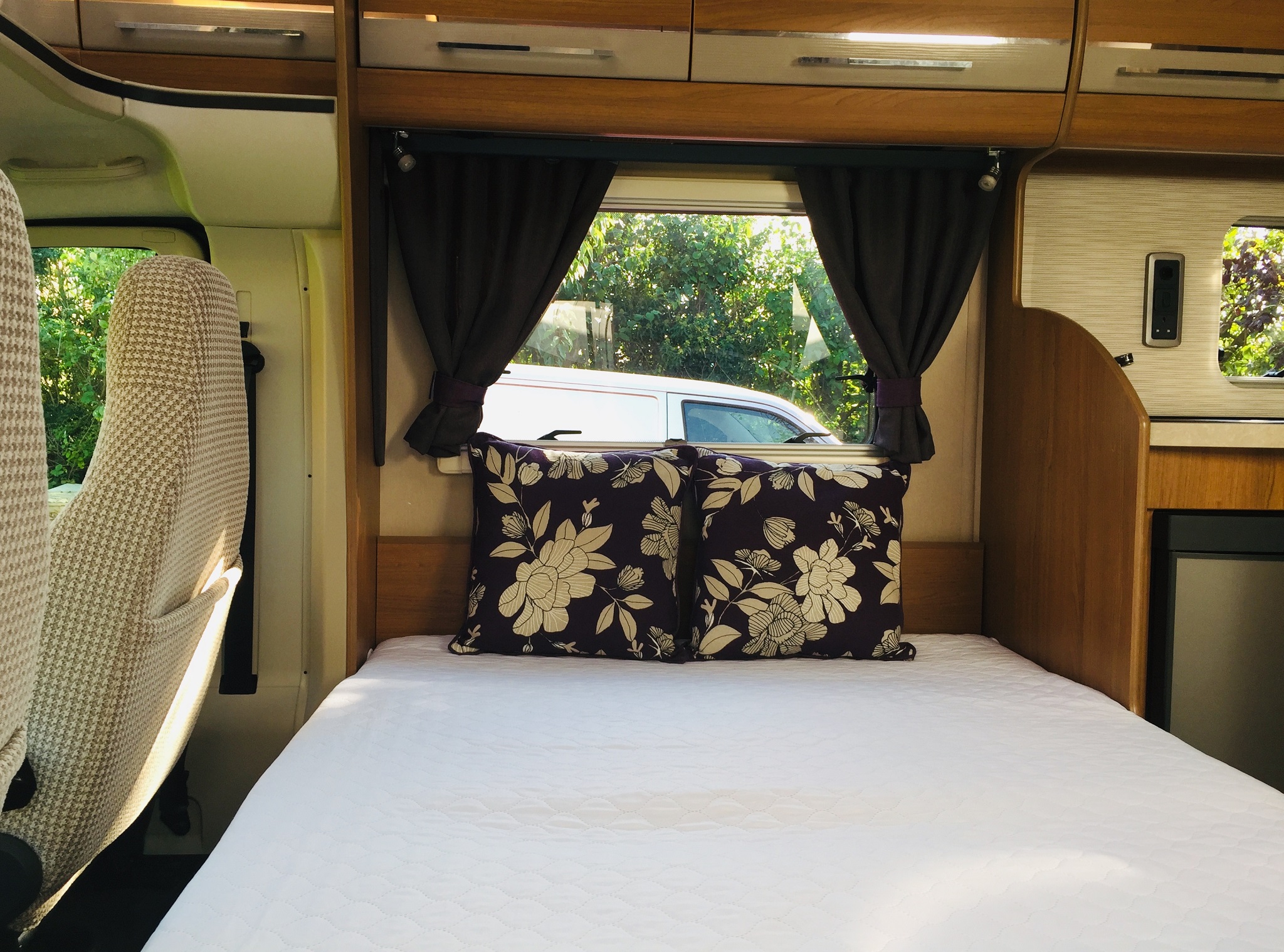 View inside an RV rental showing a neatly made bed with floral pillows and a window revealing trees and a white van.. Auto Trail Tracker RS 2016