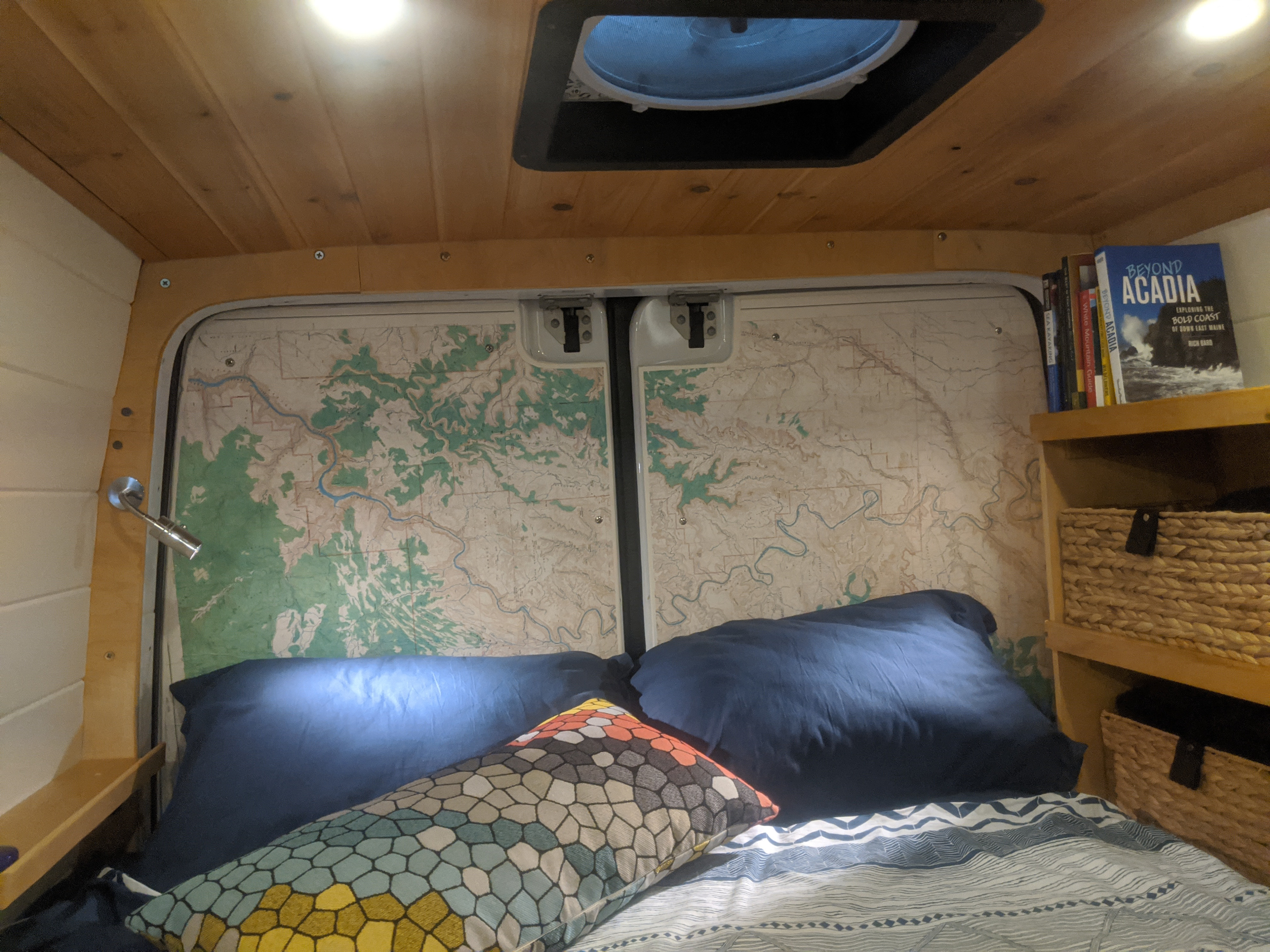 Cozy RV rental bedroom with maps on the back doors, a comfy bed, wooden ceiling, and travel books and storage baskets nearby.. Mercedes-Benz Sprinter 2012