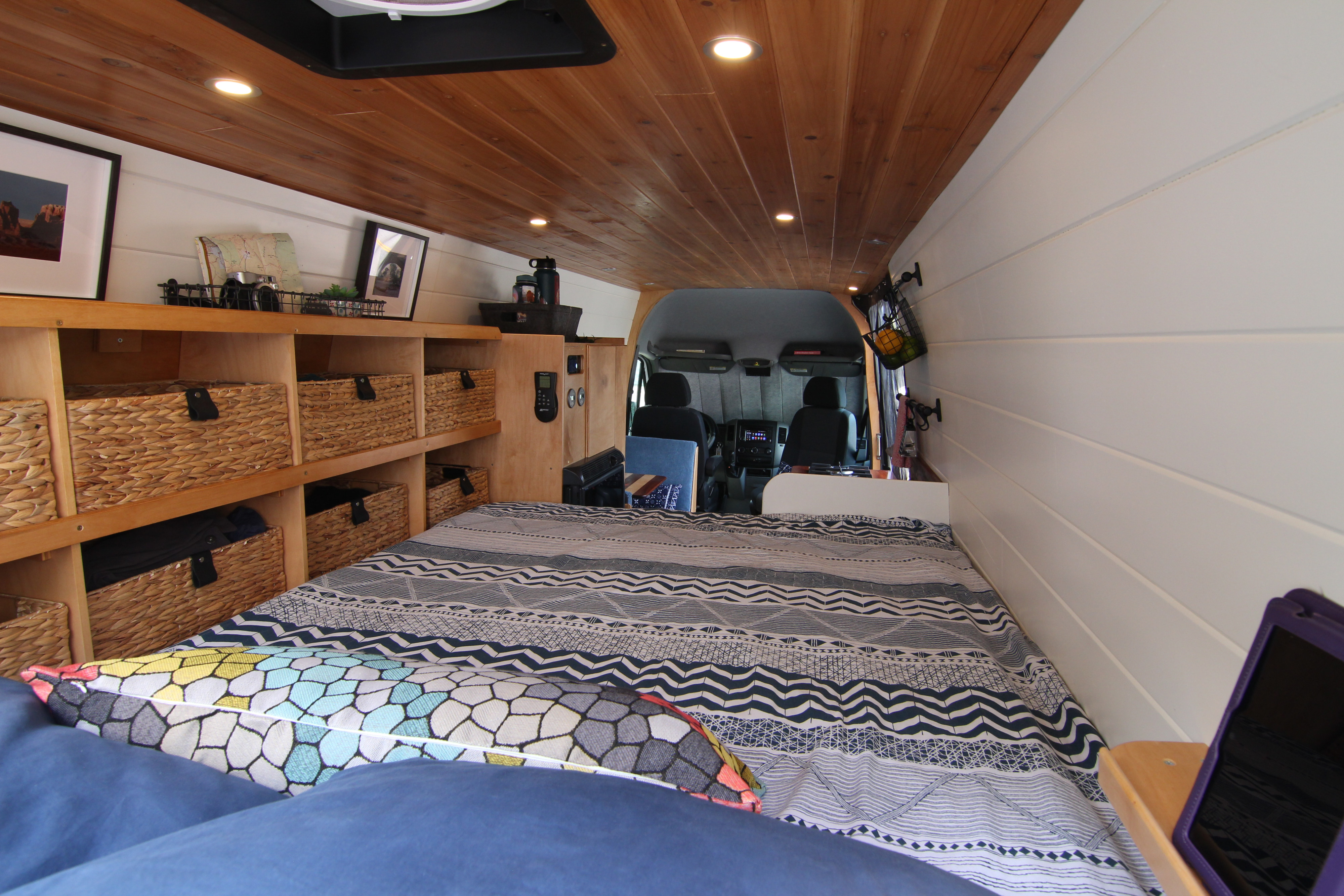 Inside a stylish RV rental, showing the bed, wood ceiling, white walls, and built-in storage with wicker baskets.. Mercedes-Benz Sprinter 2012