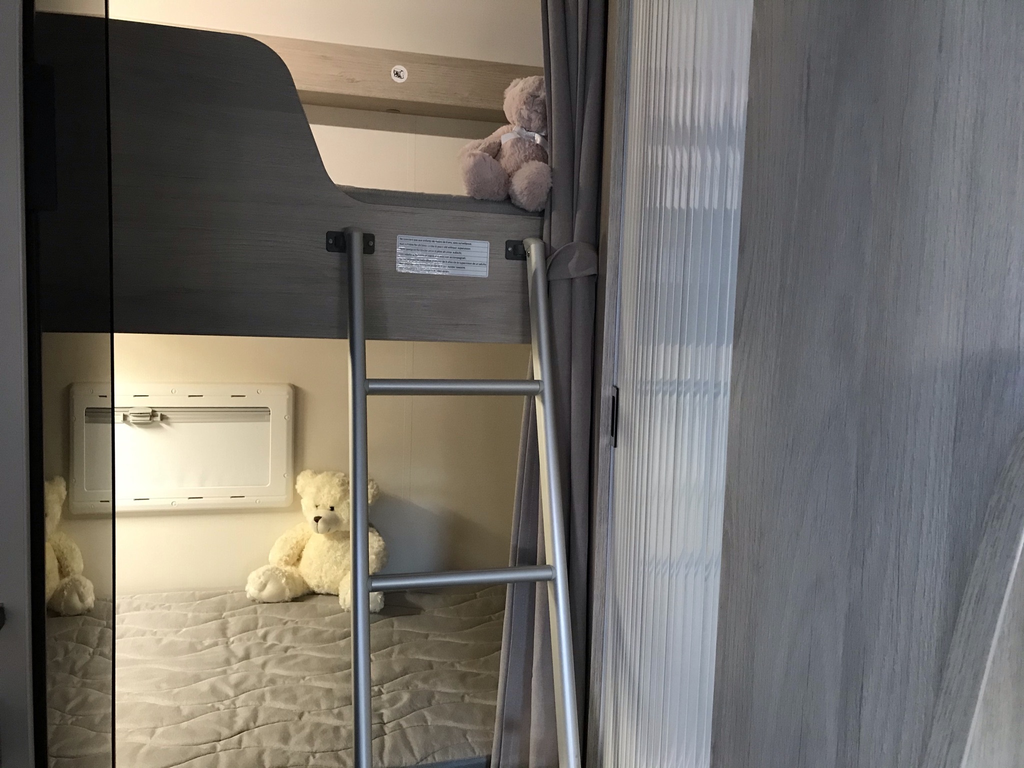 This RV rental features cozy gray bunk beds, a ladder, and two teddy bears. A bright window is visible above the bottom bunk.. Chausson C646 2019