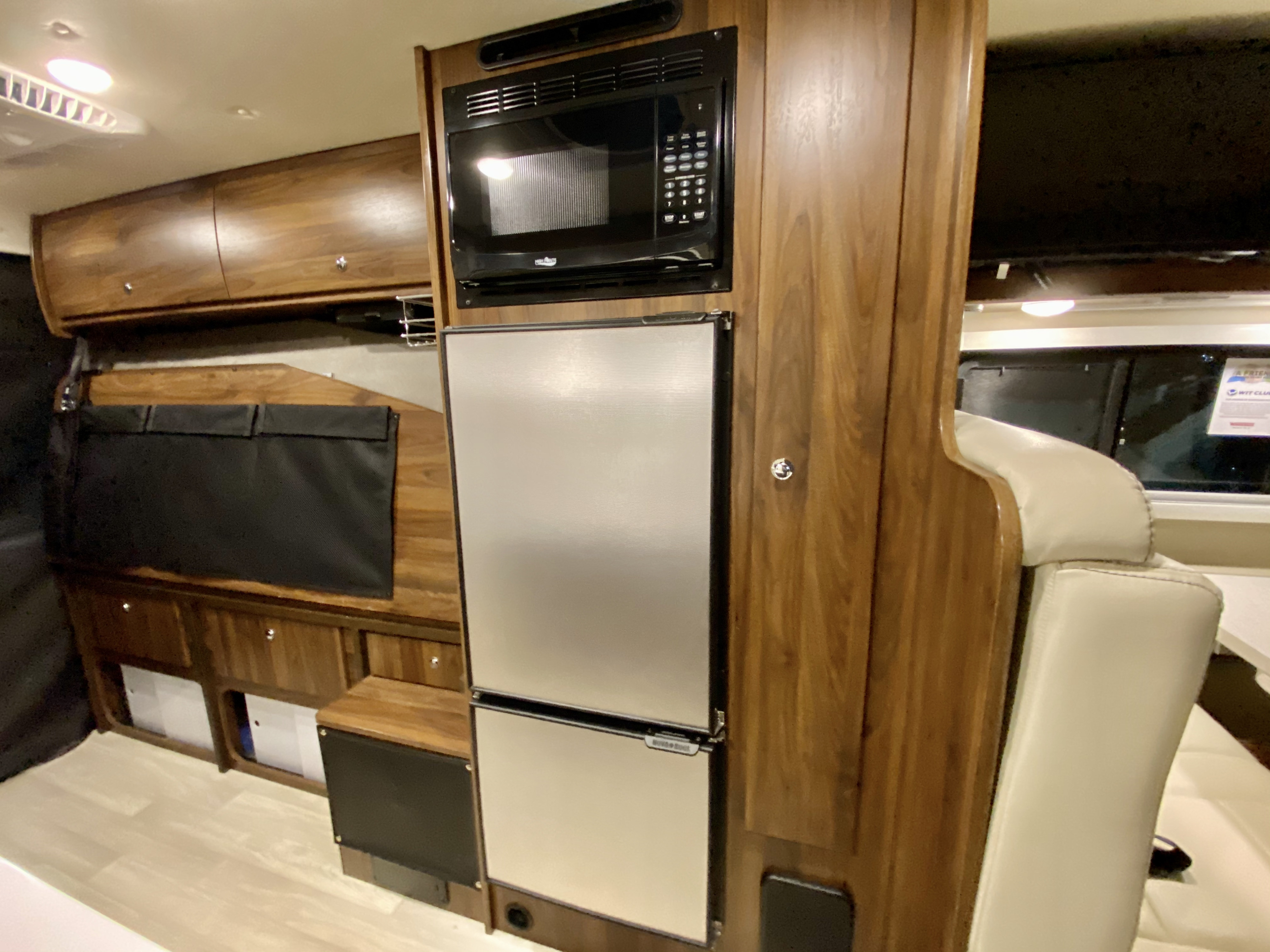 Compact RV rental kitchen with microwave, refrigerator, and rich wooden cabinetry, offering a functional space for travelers.. Winnebago Travato 2021