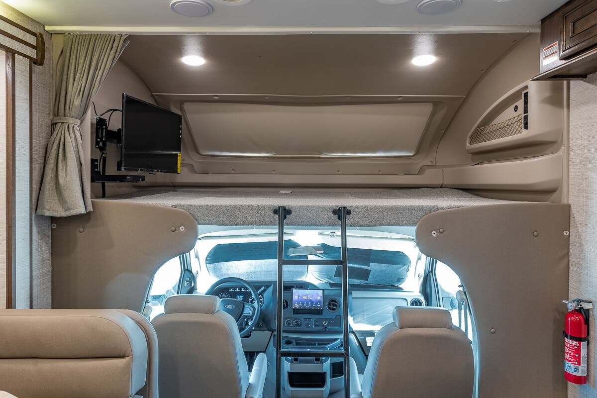 RV rental interior view: an overhead bunk with ladder, wall-mounted TV, and the driver's cab.. Entegra Coach Esteem 2021
