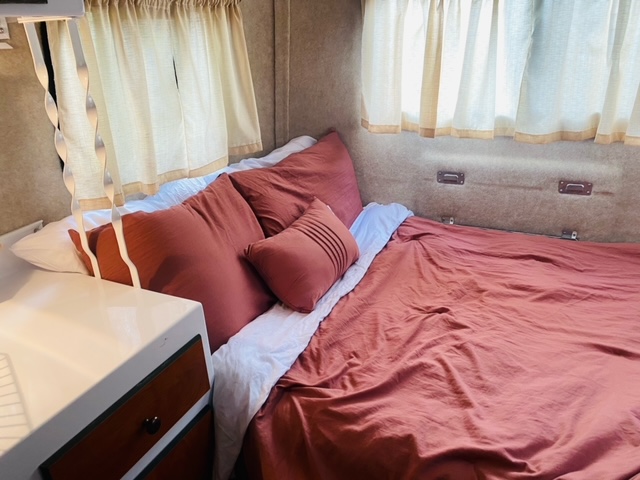 Cozy RV rental bedroom features a comfy bed with warm bedding, white sheets, pillows, curtains, and a small cabinet.. Scamp 16' 2018