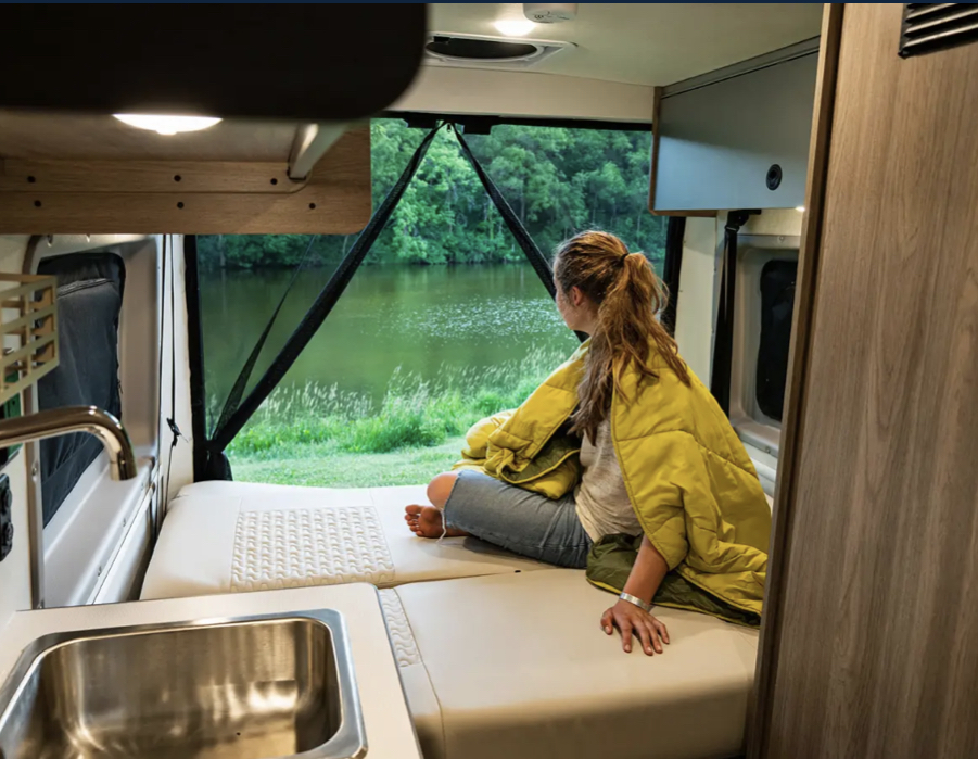 Cozy RV rental interior: A person relaxes on a bed, admiring a peaceful lake view through the large window, complete with a sink.. Winnebago Winnebago 2022