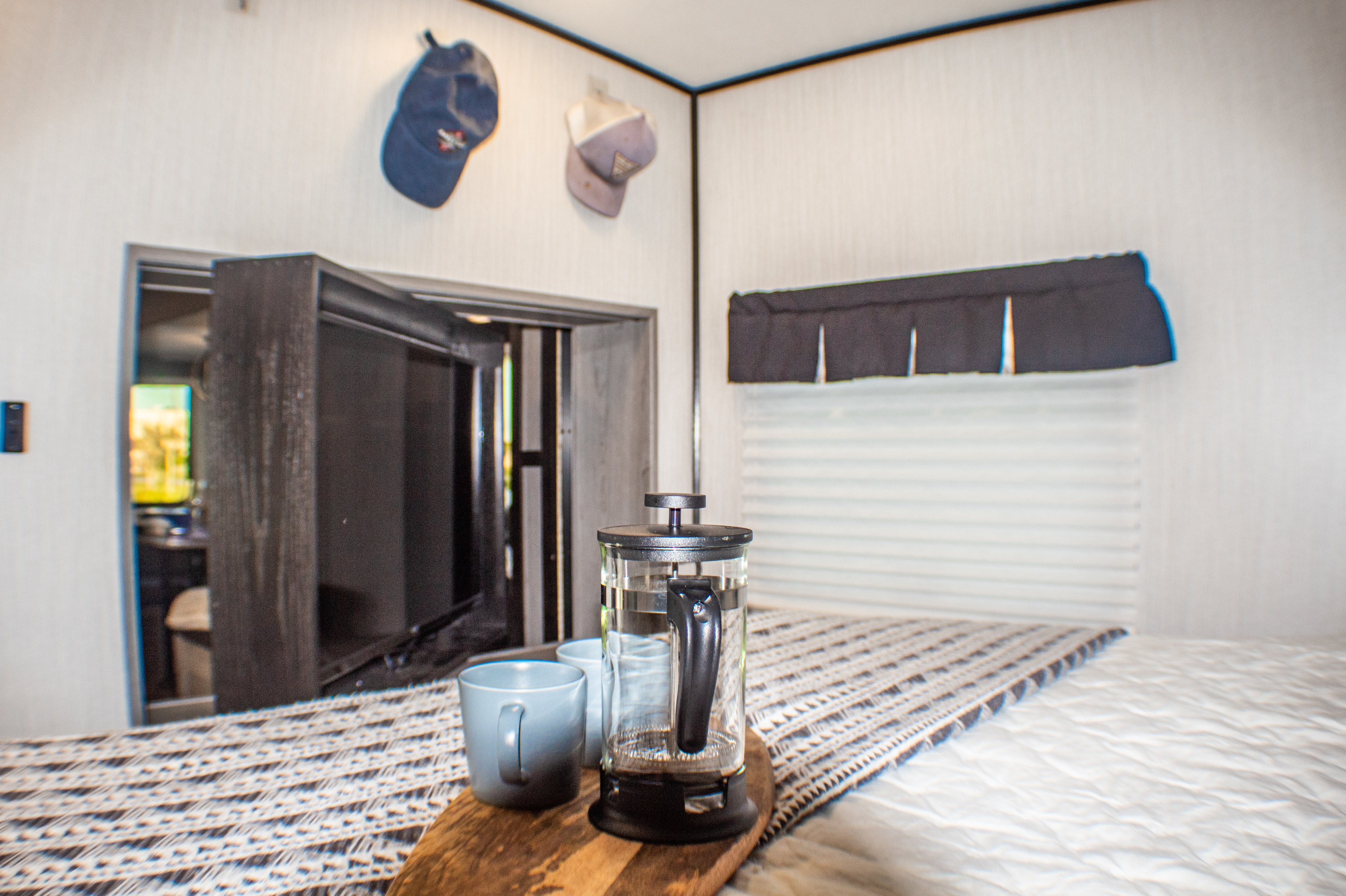 Cozy RV rental bedroom with a French press & mugs on the bed, hats on the wall, and window blinds.. Heartland M-33 2021