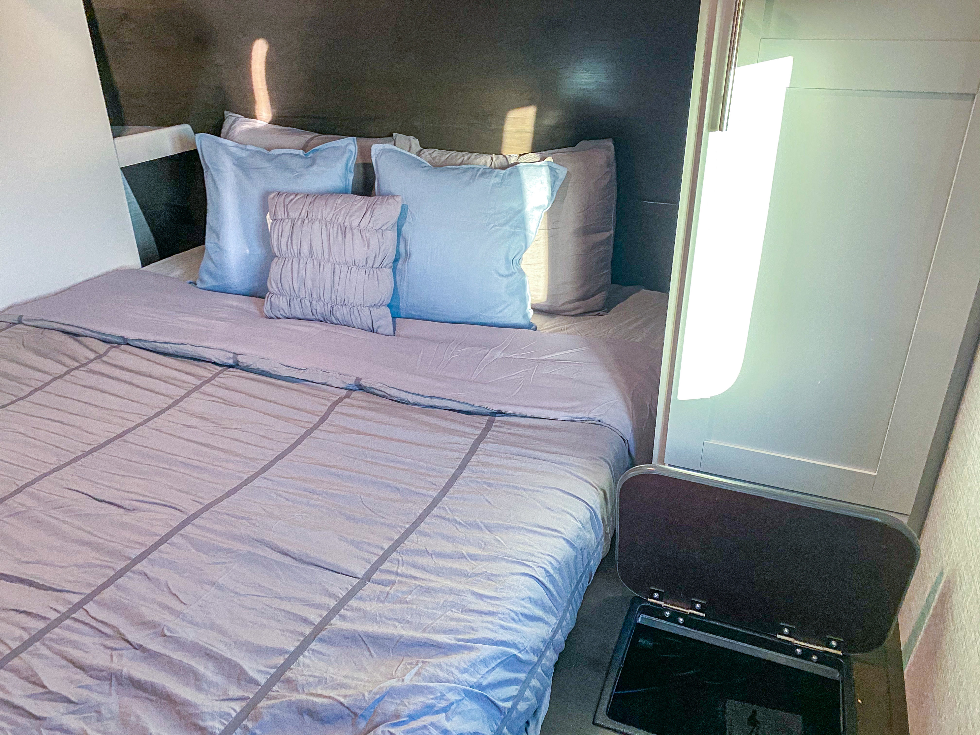 Compact RV rental sleeping area: a bed with gray and blue linens, dark headboard, and handy open storage.. Keystone Bullet 2021
