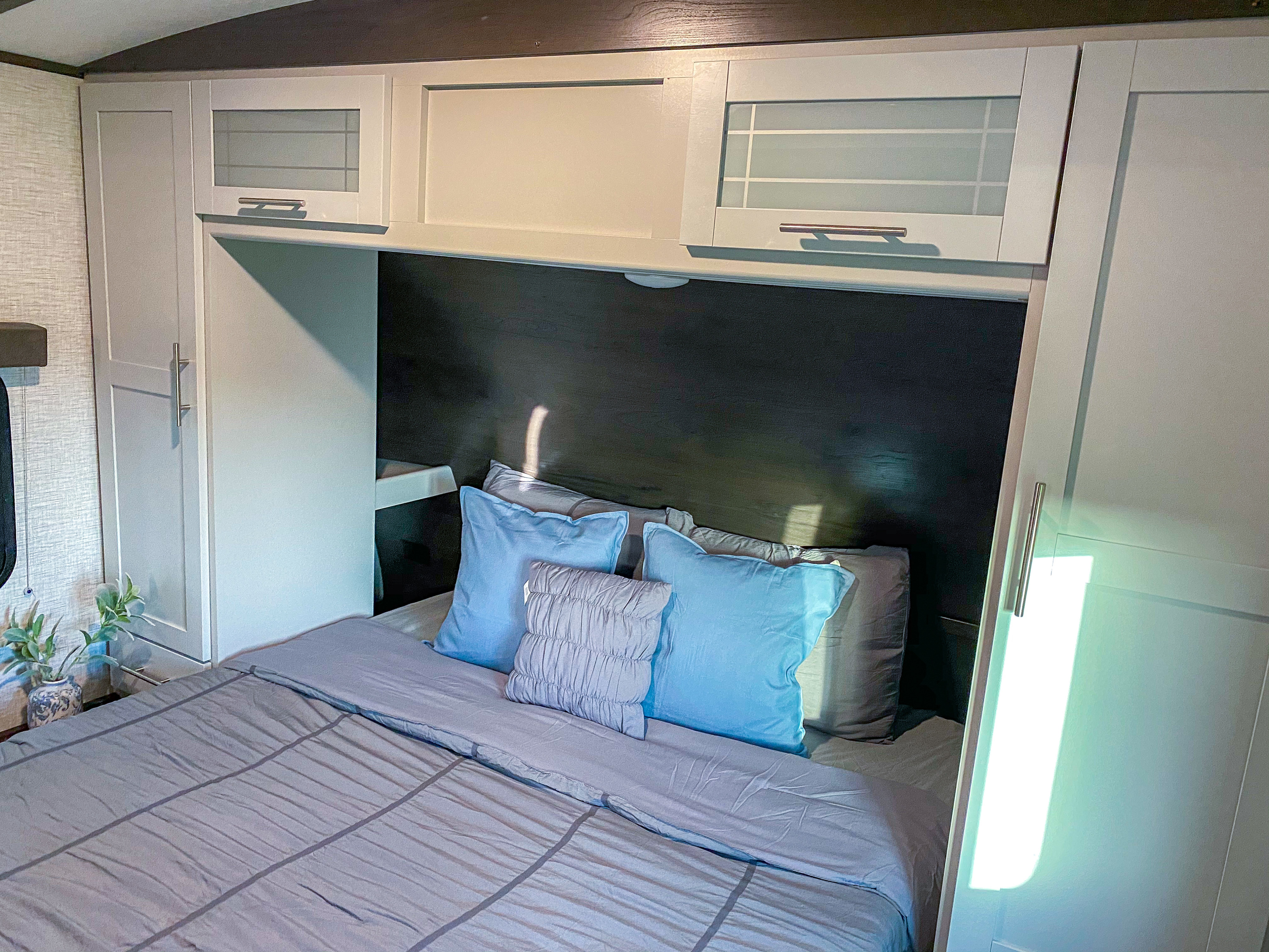 This RV rental bedroom features a gray bed, blue pillows, and white overhead cabinets, offering a compact and comfortable sleeping area.. Keystone Bullet 2021