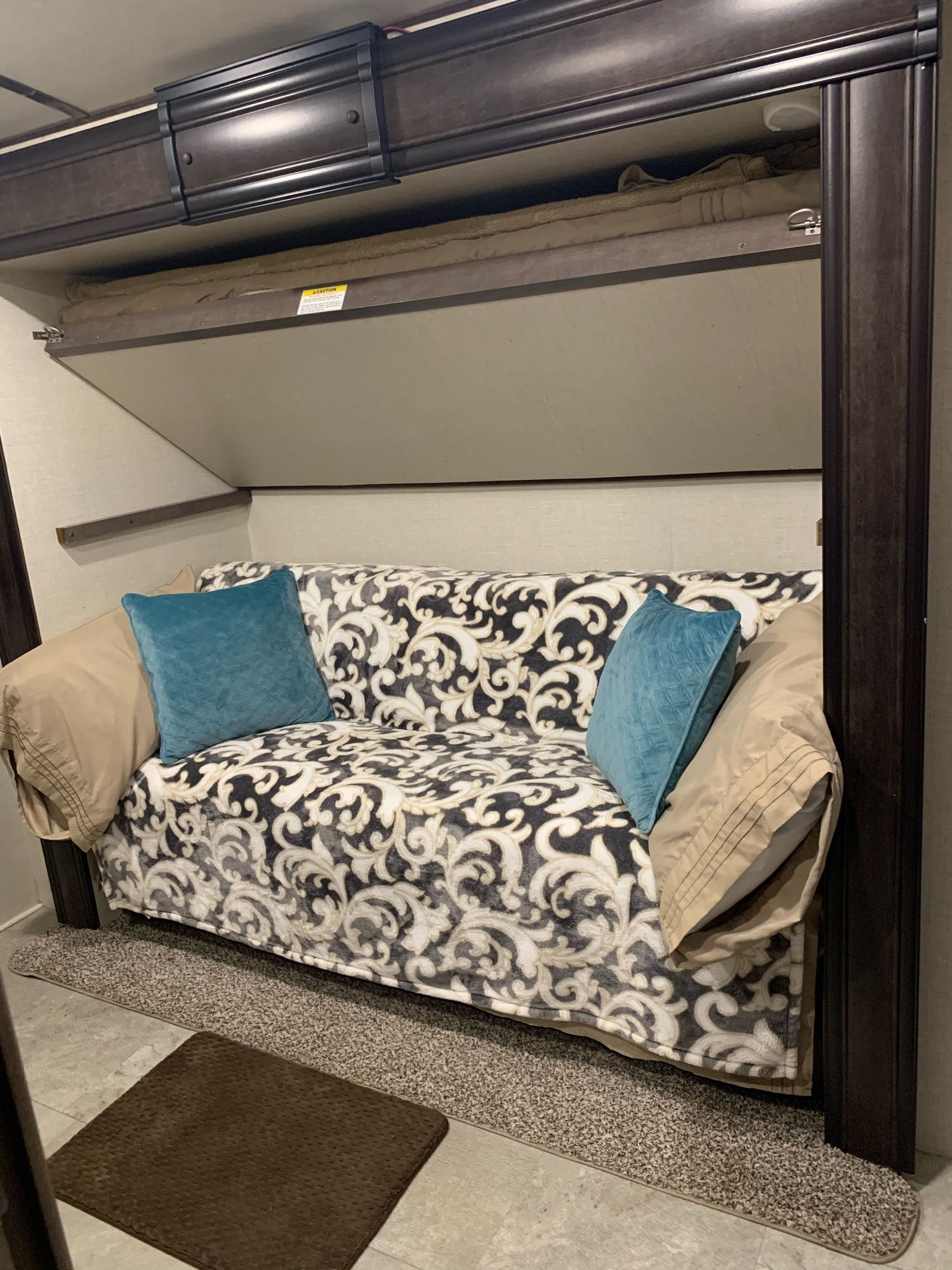 RV rental interior features a sofa with patterned throw and teal pillows, situated below a stowed upper bunk.. Crossroads Sunset Trail 2018
