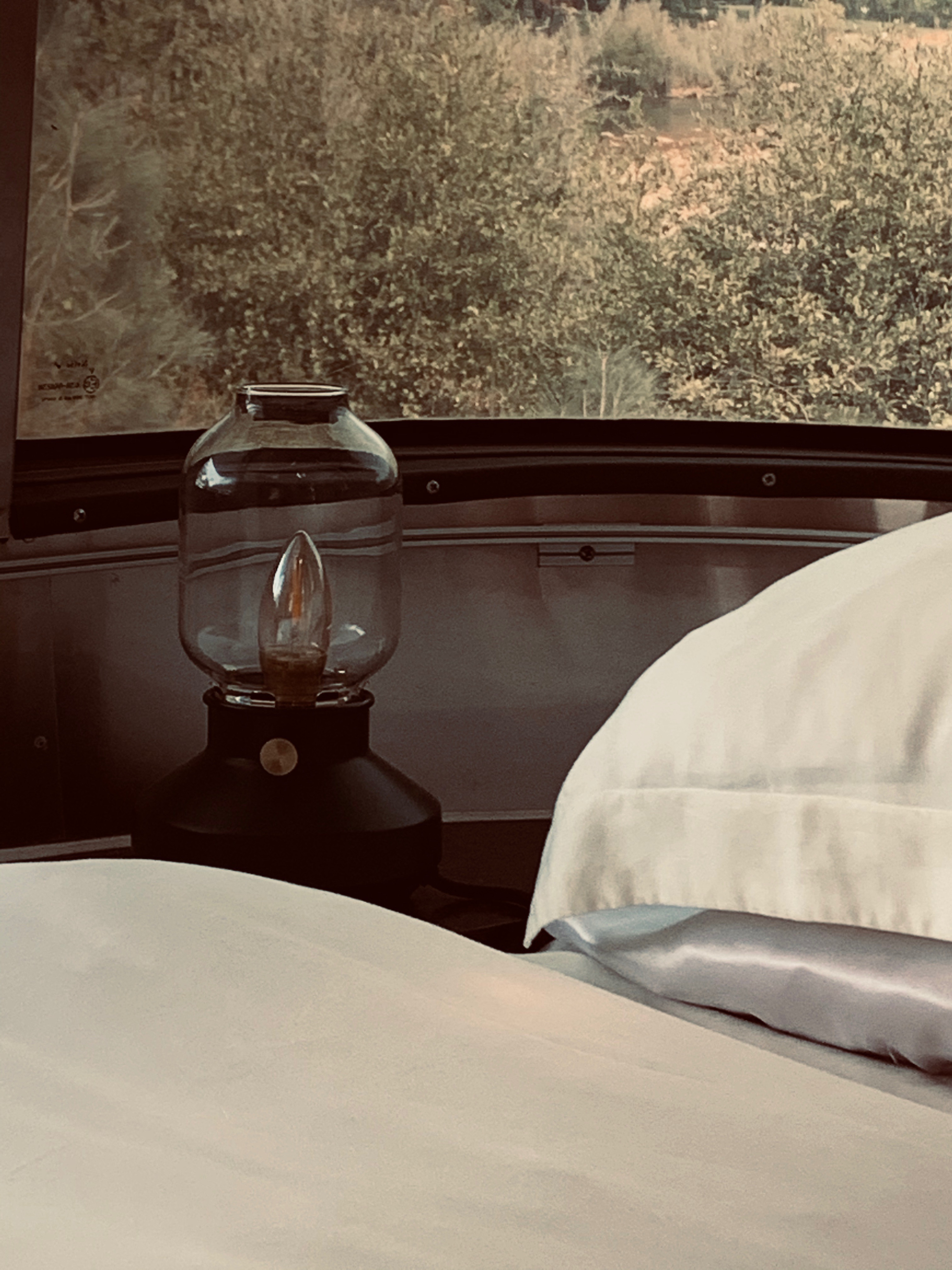 Enjoy the view from your RV rental: a cozy bed, bedside lamp, and a window overlooking nature.. Airstream Other 2011