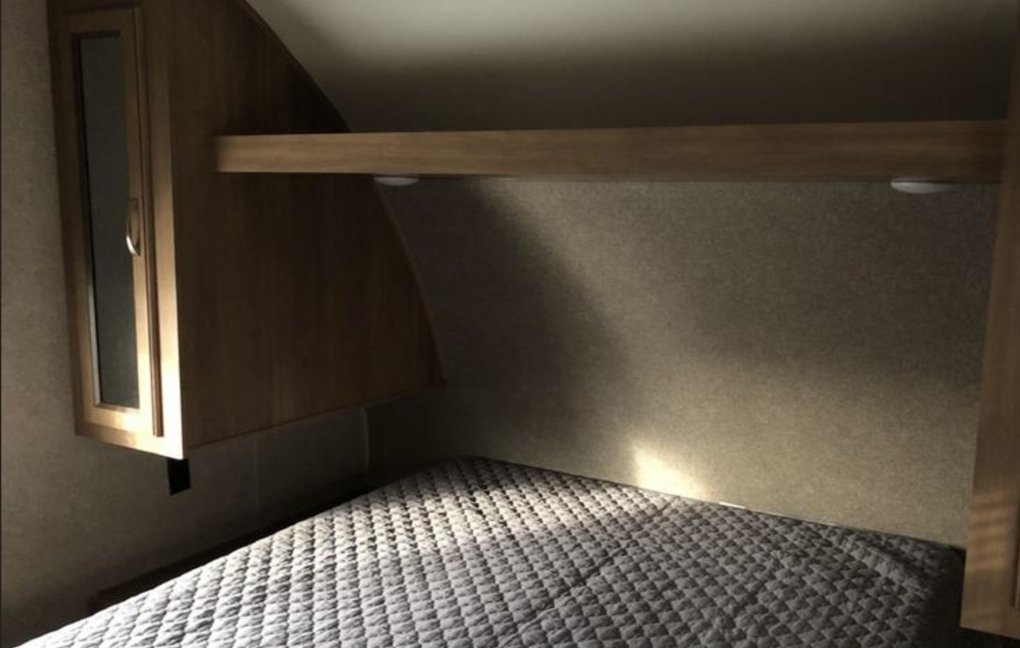 Cozy RV rental bedroom featuring a quilted bed, overhead shelf with lights, and a storage cabinet on the left.. Coachmen Catalina 261 2019