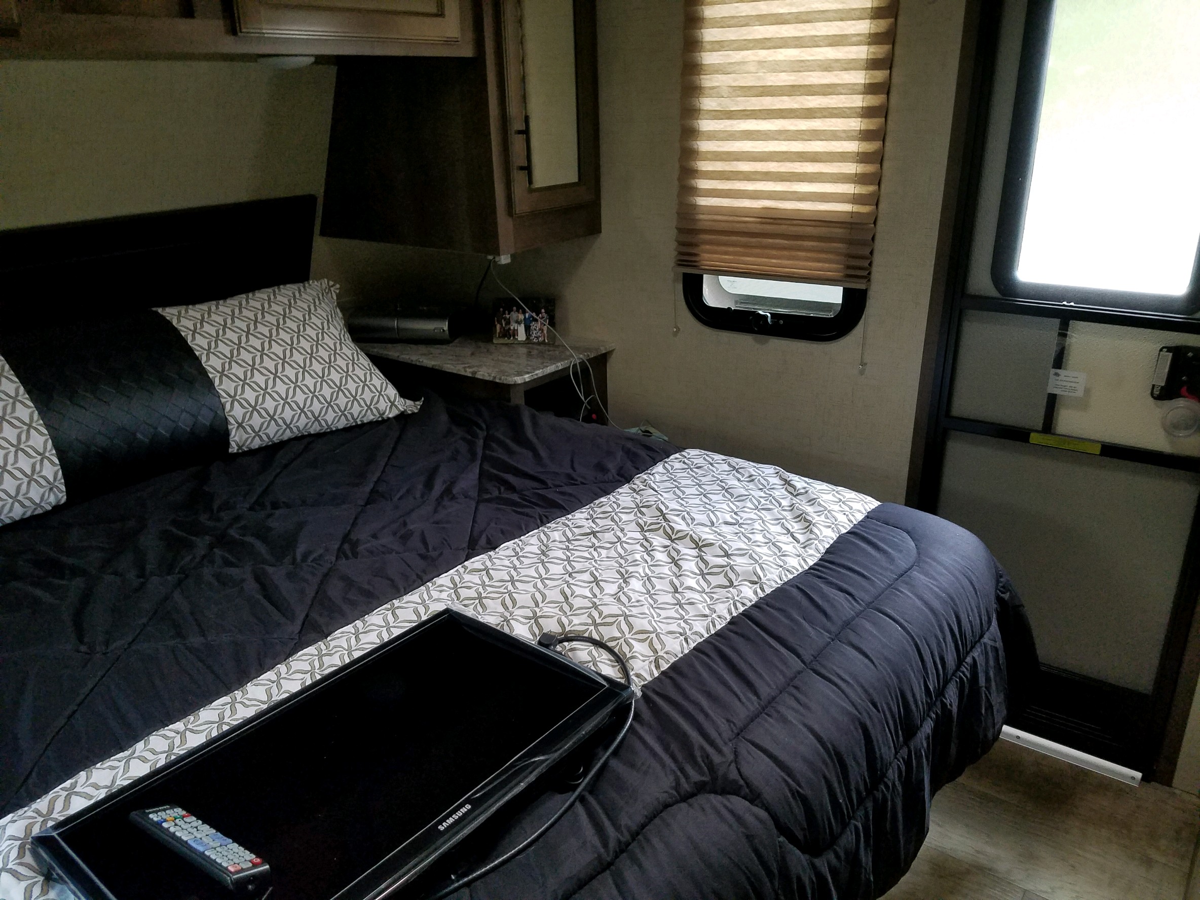RV rental bedroom featuring a bed with black and patterned bedding, a small TV, window with blinds, and an exit door.. K-Z Spree S-261RKS 2018