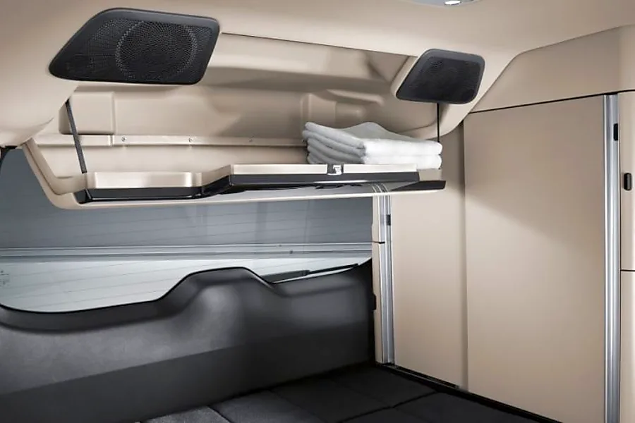 RV rental overhead storage is open, showing folded towels. Speakers and a sleeping area are visible in this clean cabin interior.. Mercedes Marco Polo Class V 250d 2022