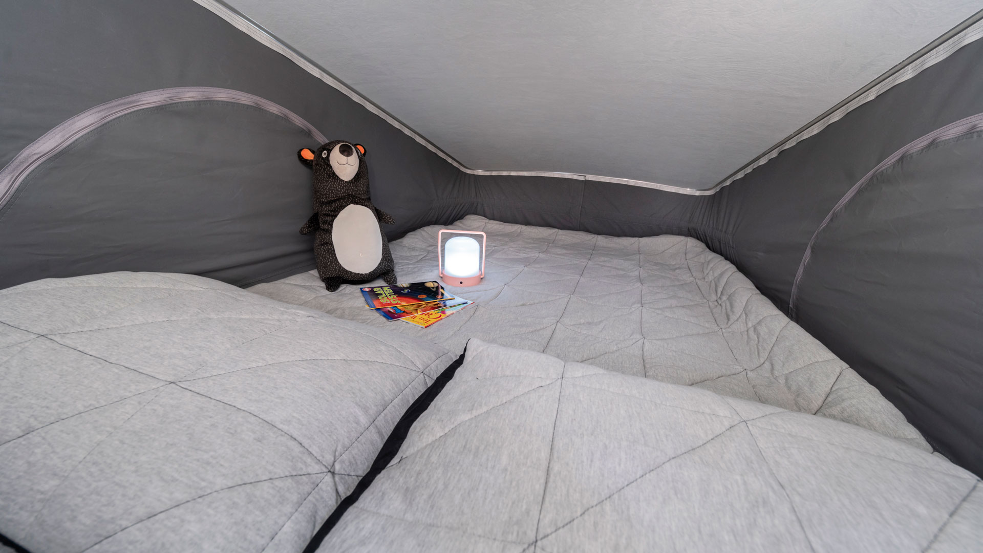 Cozy pop-top RV rental interior: a comfy bed with a bear, books, and lamp, perfect for camping adventures.. Winnebago Solis 2021