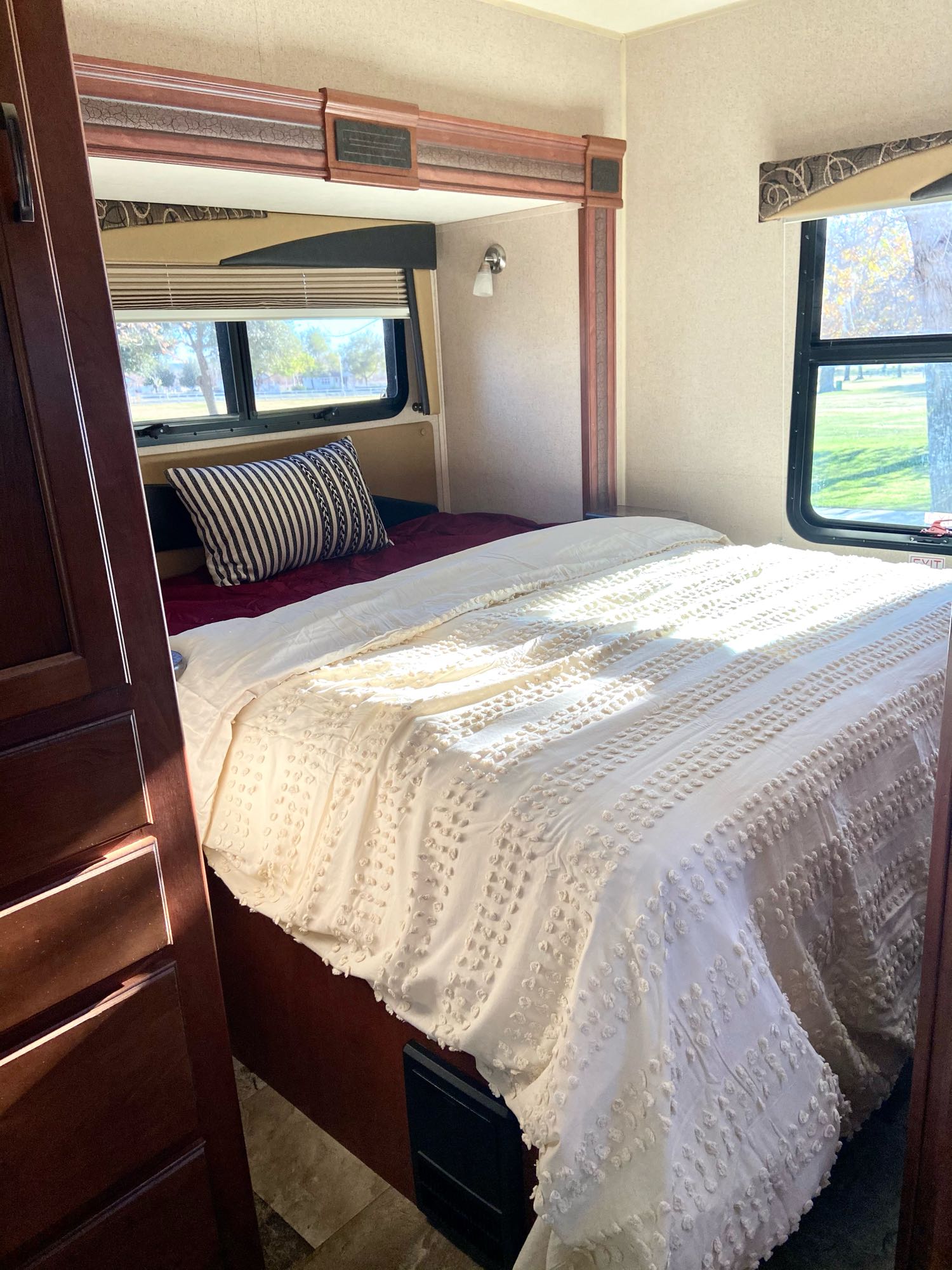 Cozy RV rental bedroom featuring a comfy bed with a white comforter, striped pillow, and sunlit windows showing outdoor views.. Jayco Redhawk 2015