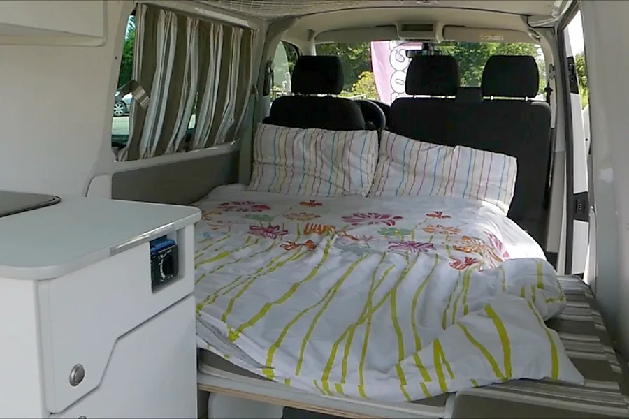 RV rental interior: a comfy bed with floral & striped bedding, filling a camper van, beside a compact kitchen.. Renault TRAFIC 2021