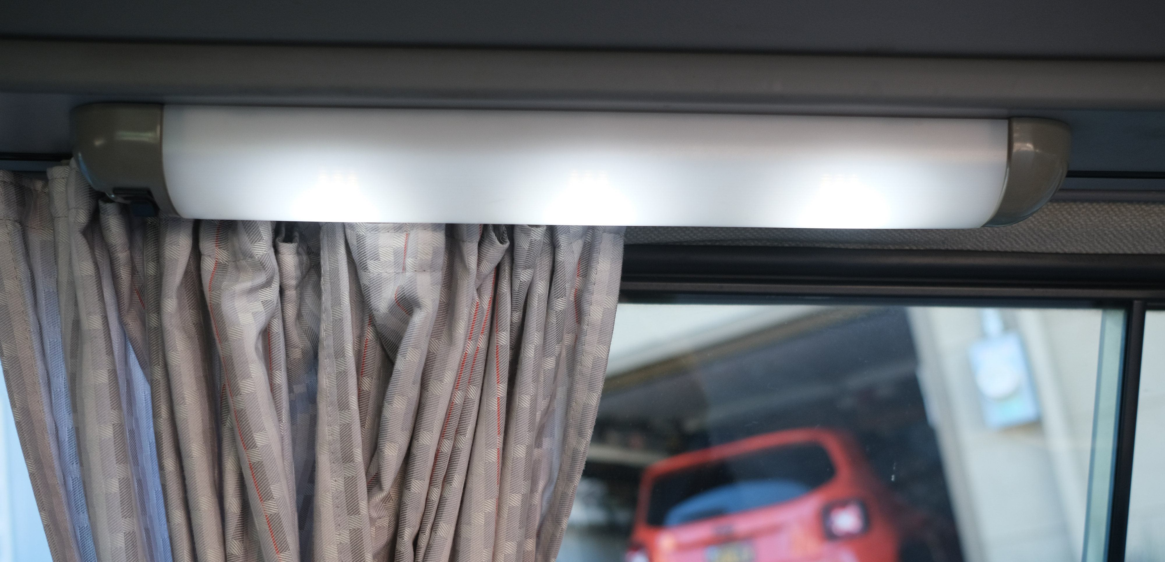 Inside an RV rental: a bright overhead light illuminates patterned curtains, with a blurry red car visible through the window.. Volkswagen Westfalia 1990