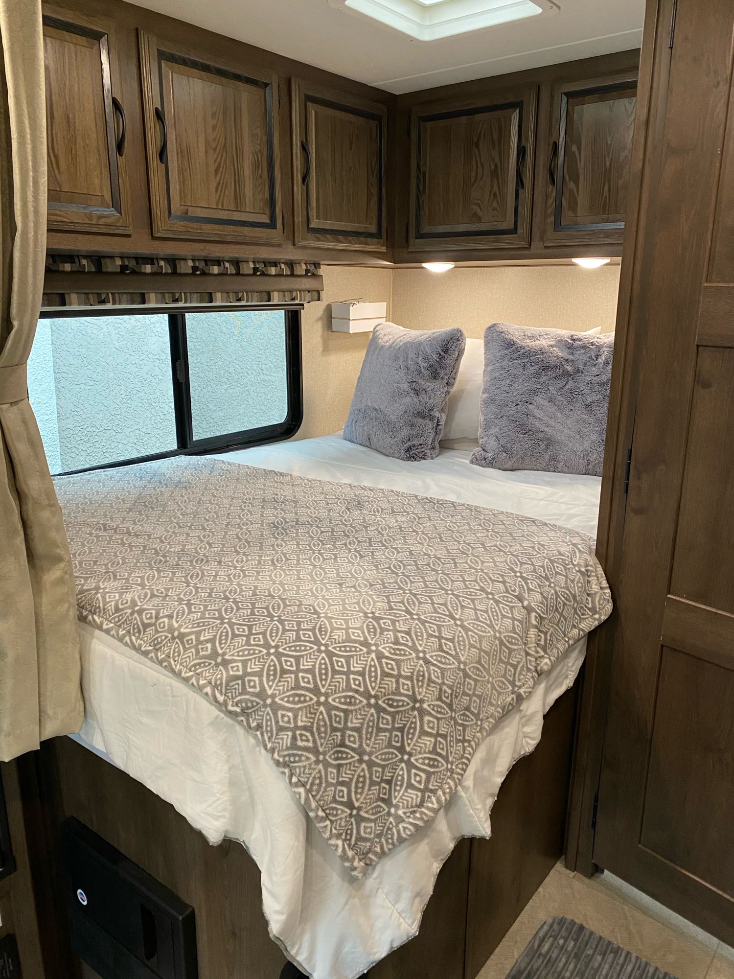 Comfortable RV rental bedroom featuring a bed with gray pillows & patterned blanket, overhead wooden cabinets, and window. Ideal for travel.. Coachmen Prism 2019