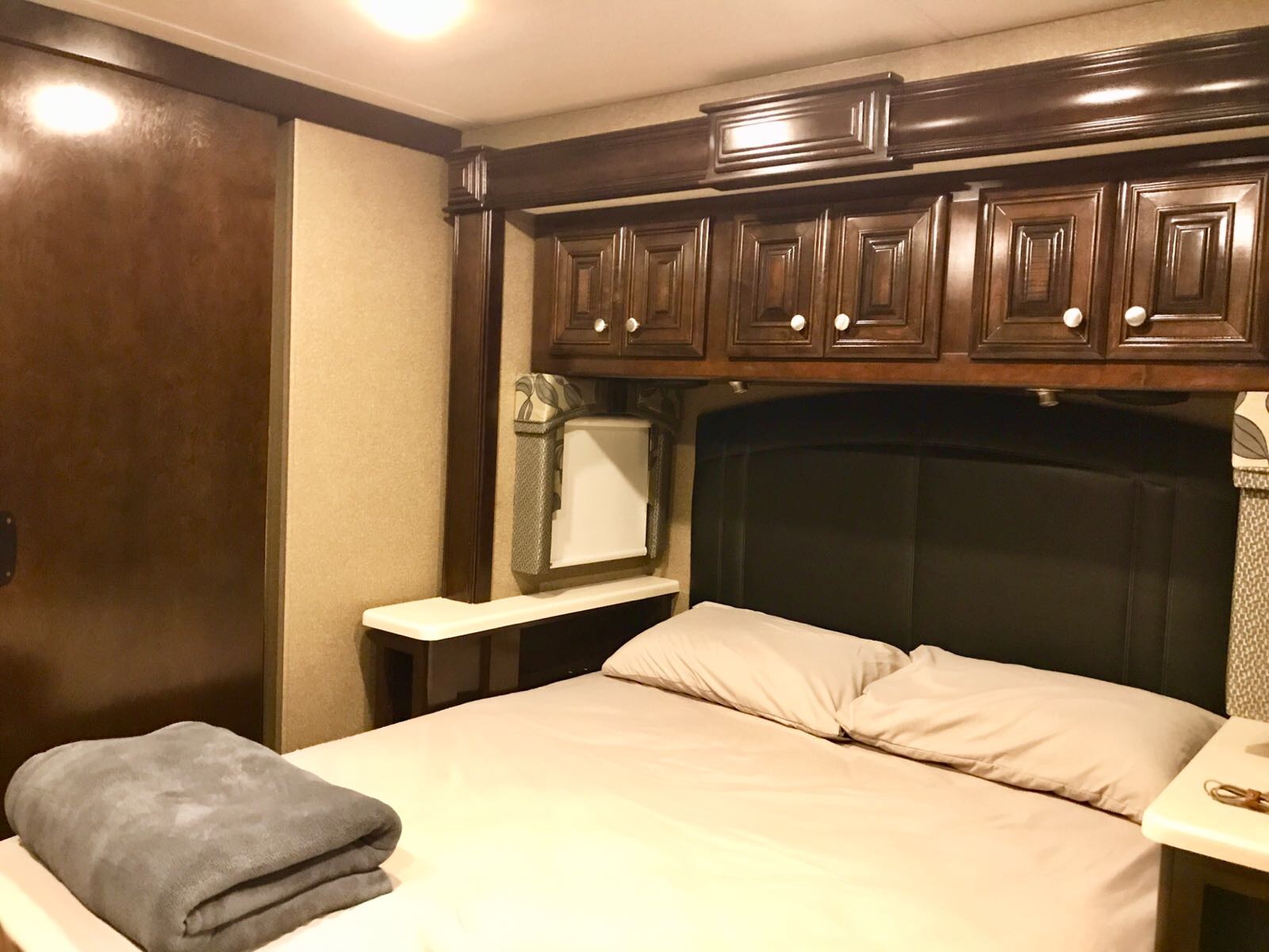 Inside an RV rental: a tidy bedroom with a queen-sized bed, dark wood overhead cabinets, and a sleek black headboard.. Thor Motor Coach Palazzo 2016