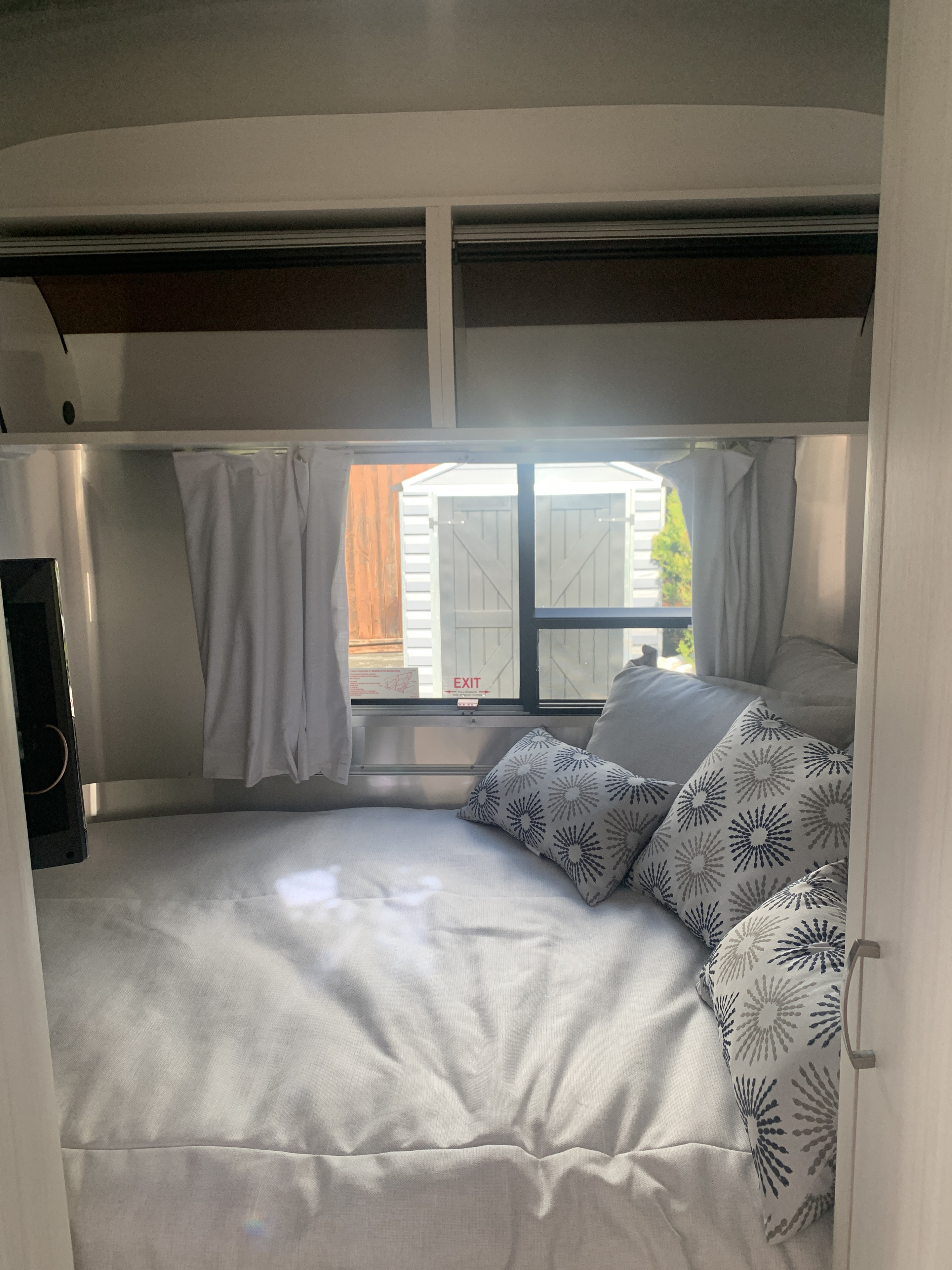 RV rental cozy sleeping area: bed with sunburst pillows, TV, overhead storage, and a bright window.. Airstream Bambi 16” 2021