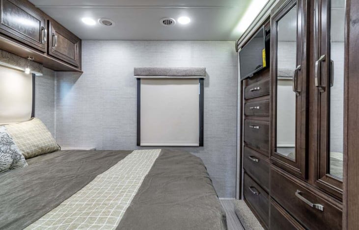 Modern RV rental bedroom features dark wood cabinets, comfortable bed, window, TV, and wardrobe.. Entegra Coach Esteem 2021