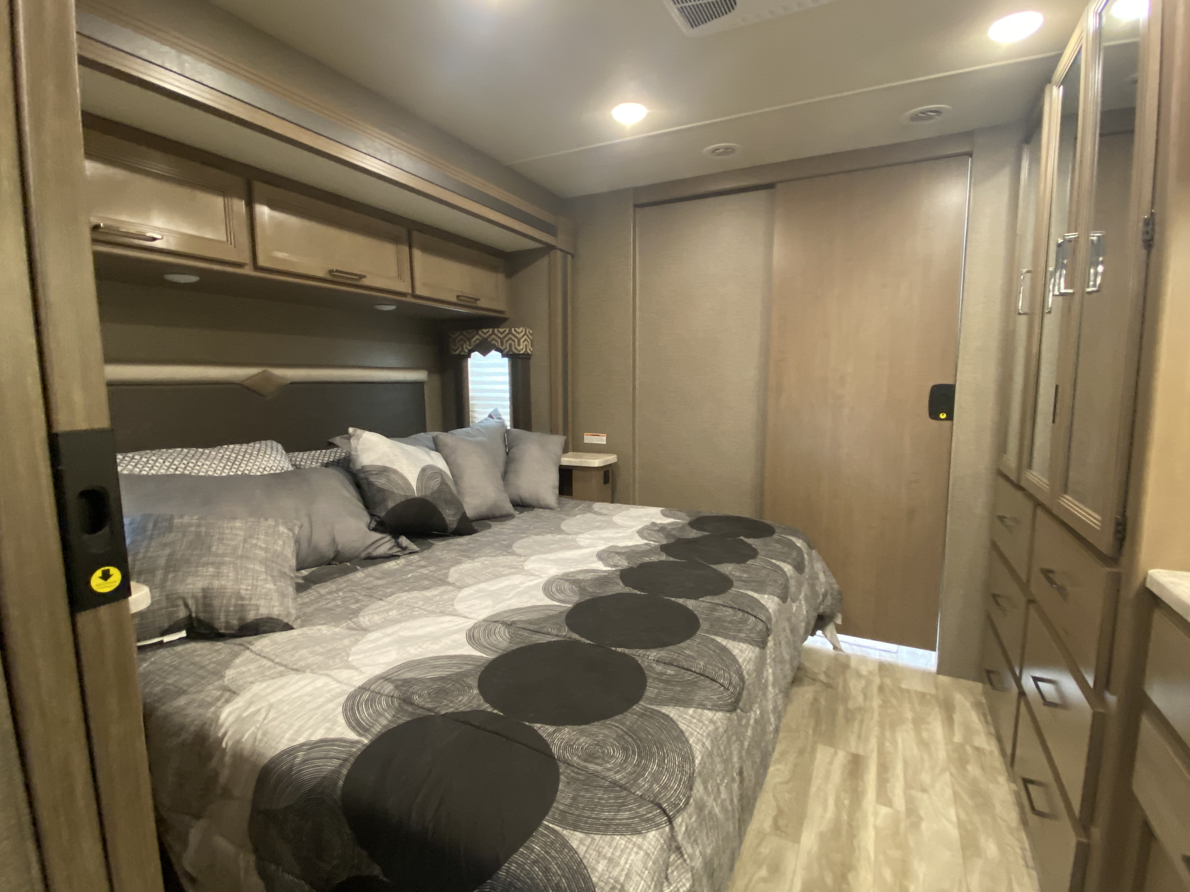 Cozy RV rental bedroom with patterned comforter, overhead storage, and mirrored wardrobe for your travels.. Thor Motor Coach Hurricane 2021