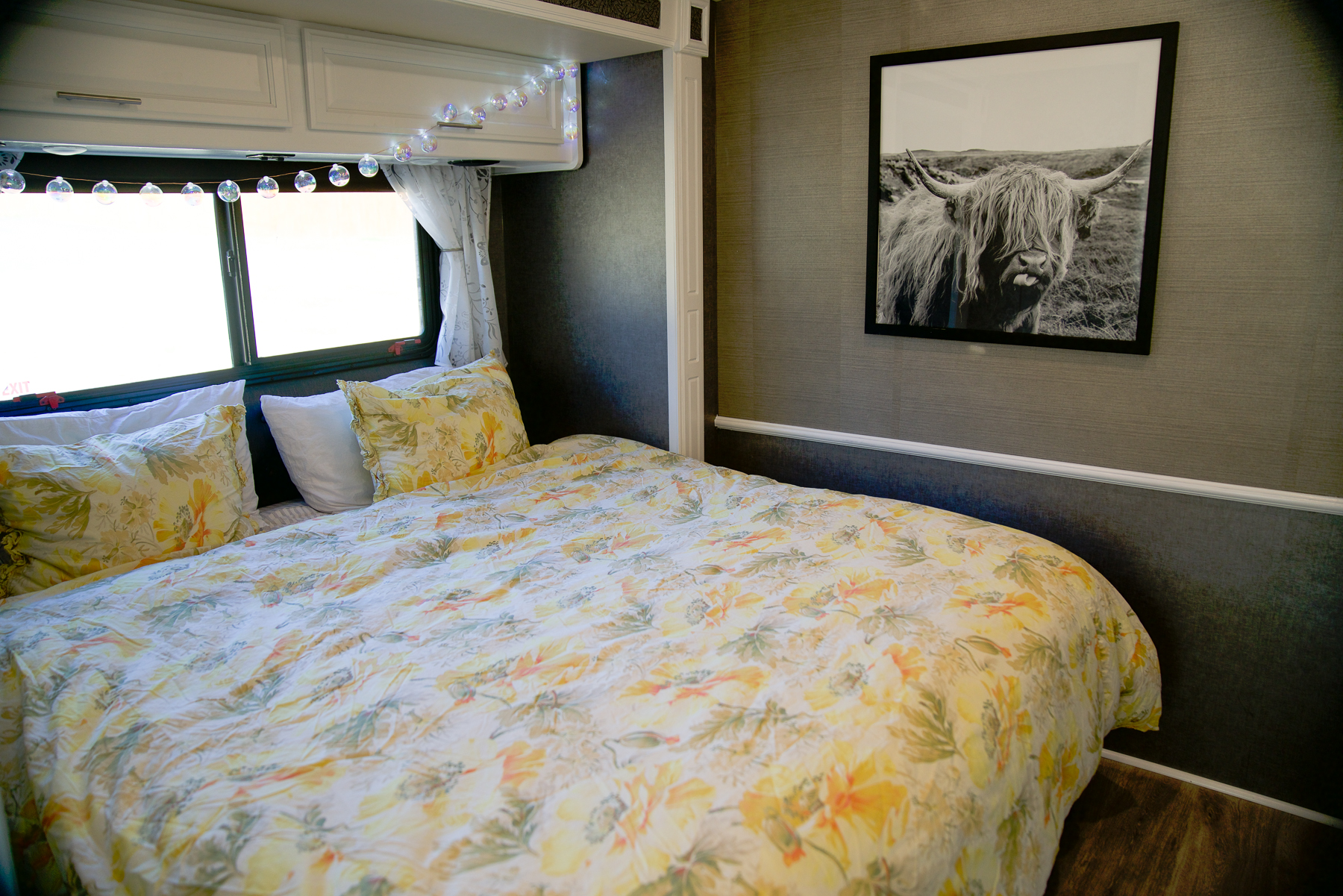 Inviting RV rental bedroom with floral bedding, string lights, bright windows, and a striking Highland cow print on gray walls.. Fleetwood Bounder 2015