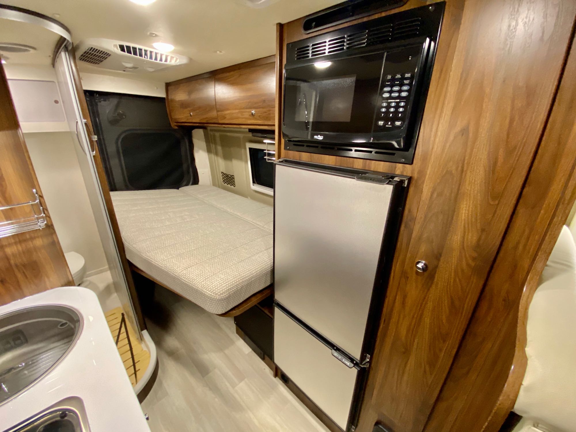 Step inside this compact RV rental! It features a bed, kitchen with sink, microwave, and fridge, all with stylish wood cabinetry.. Winnebago Travato 2021