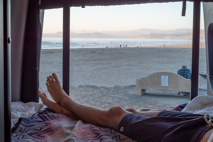 Enjoying an RV rental. Feet up, looking out the window at a sandy beach, ocean, and distant mountains. A serene view.. Winnebago Revel 2021