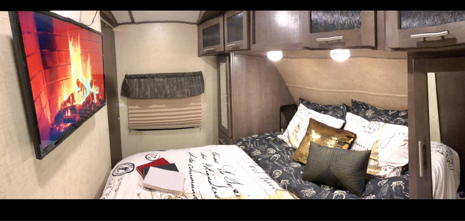 This RV rental bedroom has a mounted TV displaying a fireplace, a bed with various pillows & books, and wood cabinets/storage.. Forest River Surveyor 2016