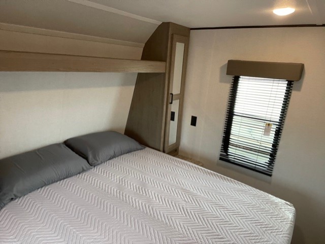 Spacious RV rental bedroom featuring a comfy bed with grey pillows, overhead bunk, wardrobe, and window with blinds.. Keystone Hideout 2021