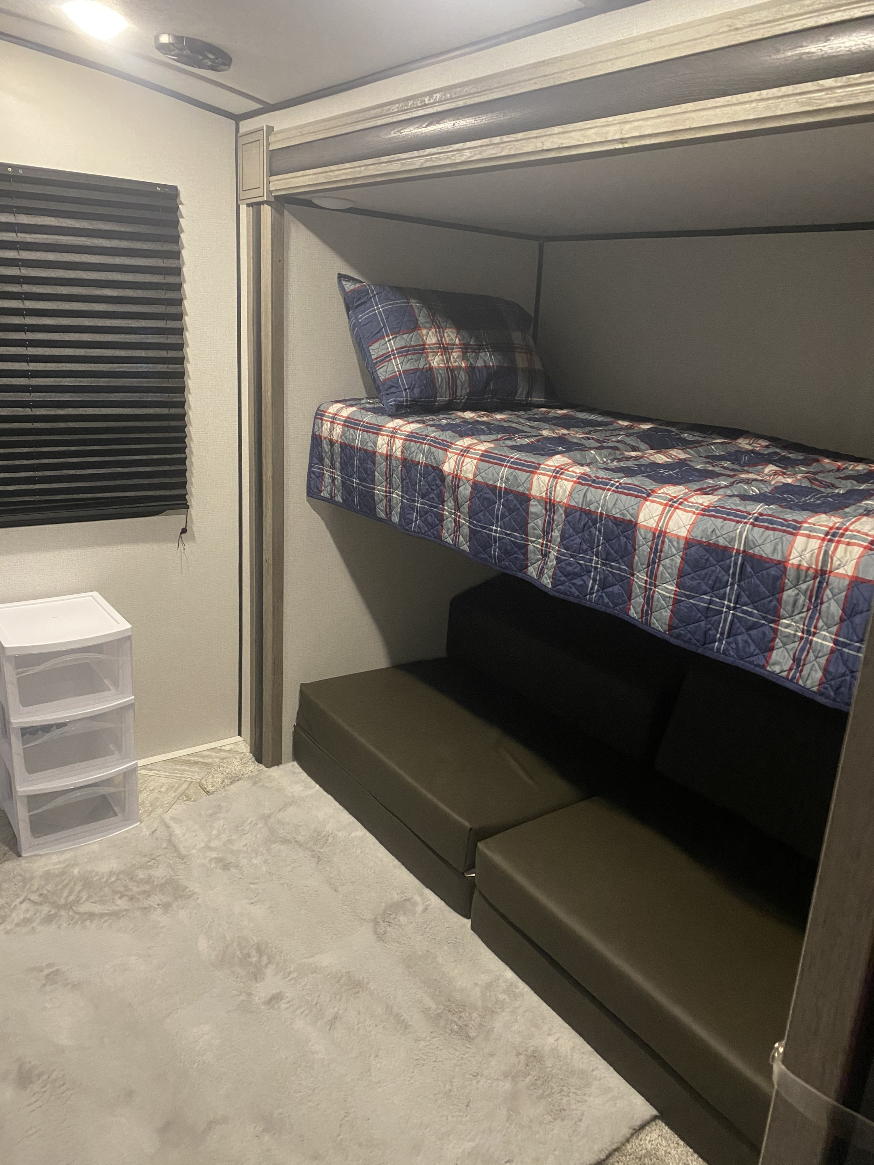RV rental bunk bed setup: plaid bedding, dark convertible cushions below, plastic drawers, and a fluffy rug.. Keystone Springdale 2021