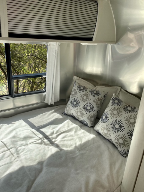 Sun-drenched RV rental bedroom: comfy bed with patterned pillows, metallic walls, and a tree-filled window view.. Airstream Bambi 16RB 2020