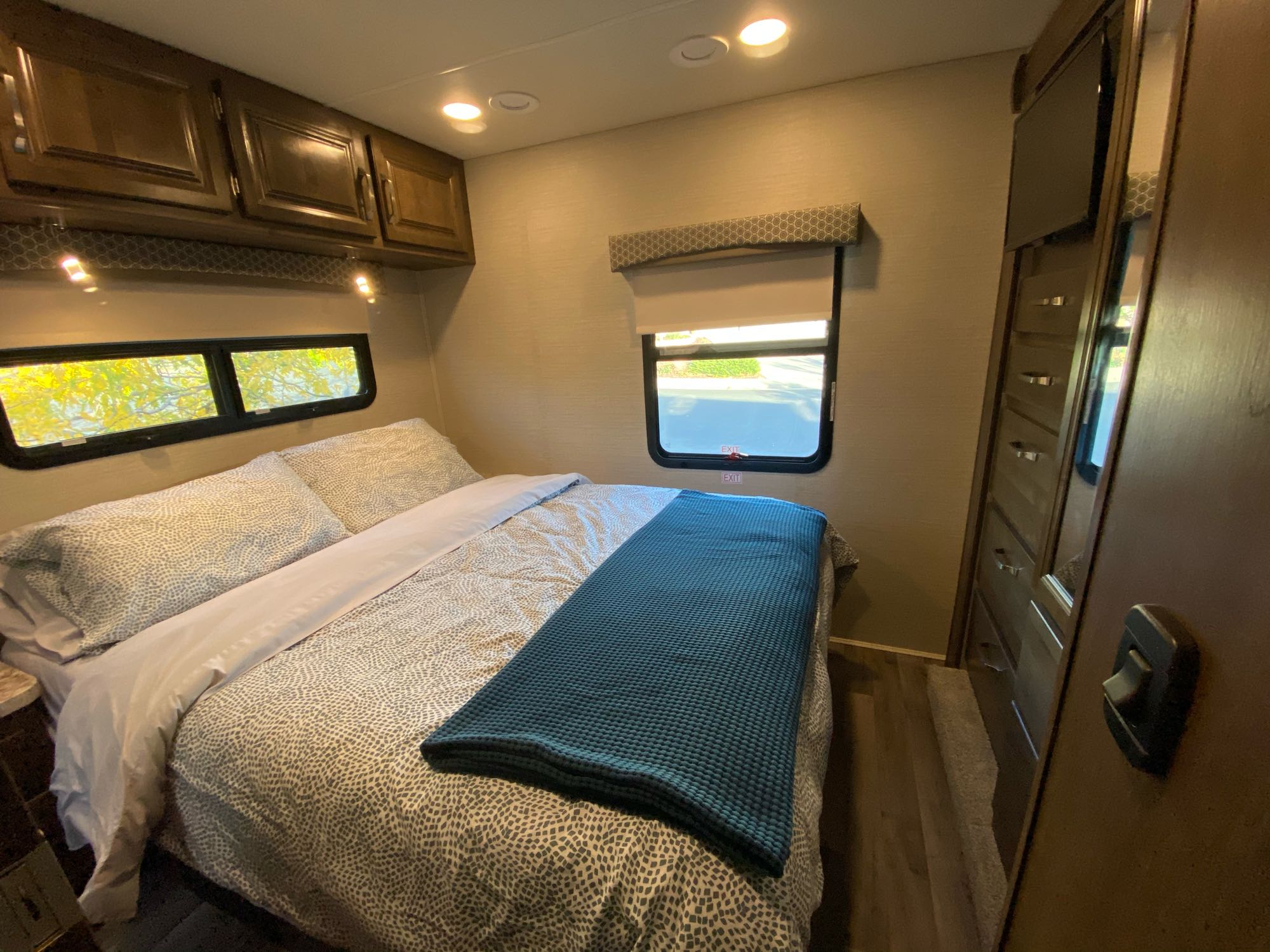 Inside an RV rental bedroom: cozy bed with patterned bedding, overhead cabinets, windows, ceiling lights, and TV/storage.. Jayco Greyhawk 2019