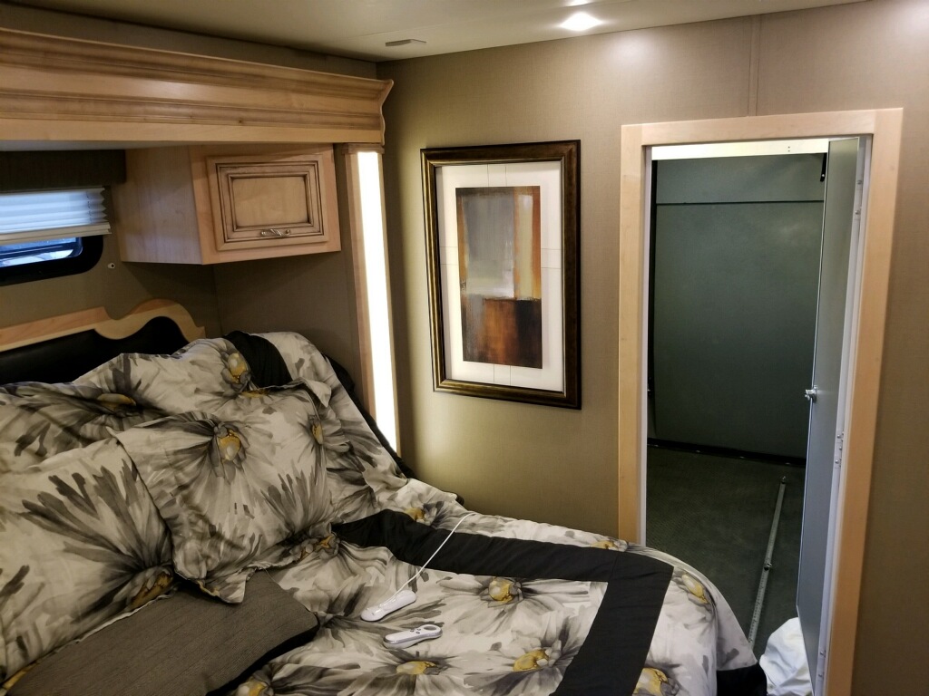 Cozy RV rental bedroom: floral bedding, overhead cabinet, and controls on the bed. Open door to an adjacent space for travel.. Newmar Canyon Star 2015