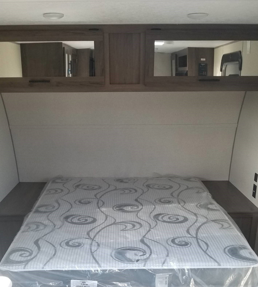 Inside an RV rental: a compact bedroom with a new mattress in plastic and reflective overhead cabinets.. Prime Time Avenger 16FQ 2021